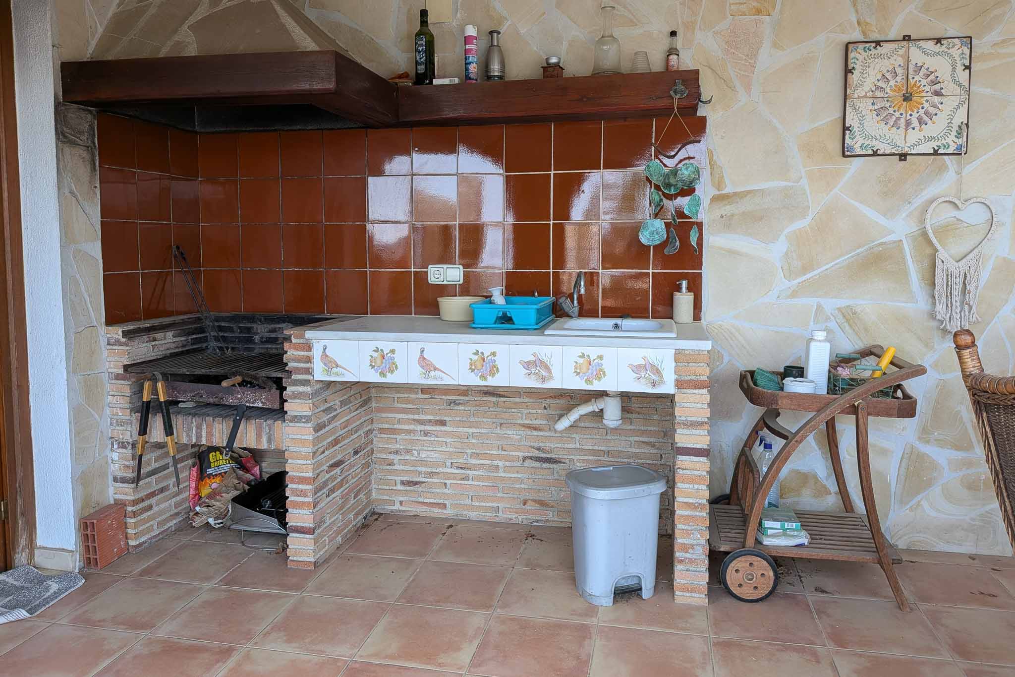 Cuisine d'été extérieure avec barbecue en briques, évier intégré et plan de travail carrelé, le tout adossé à un mur en pierre naturelle apparente.