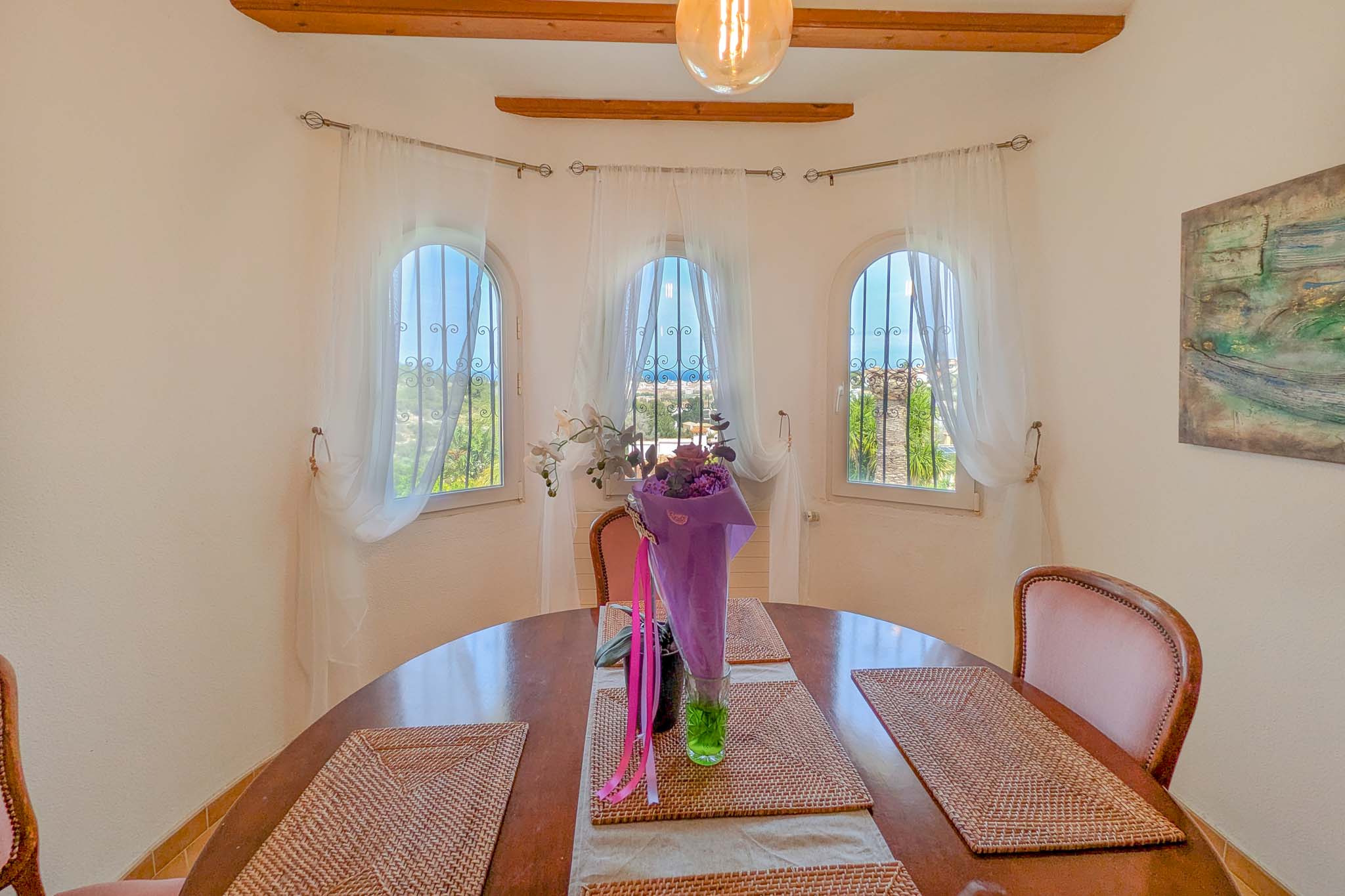 Espace salle à manger avec table ronde en bois et trois fenêtres en plein cintre vue mer. La pièce se distingue par ses poutres apparentes et sa luminosité.