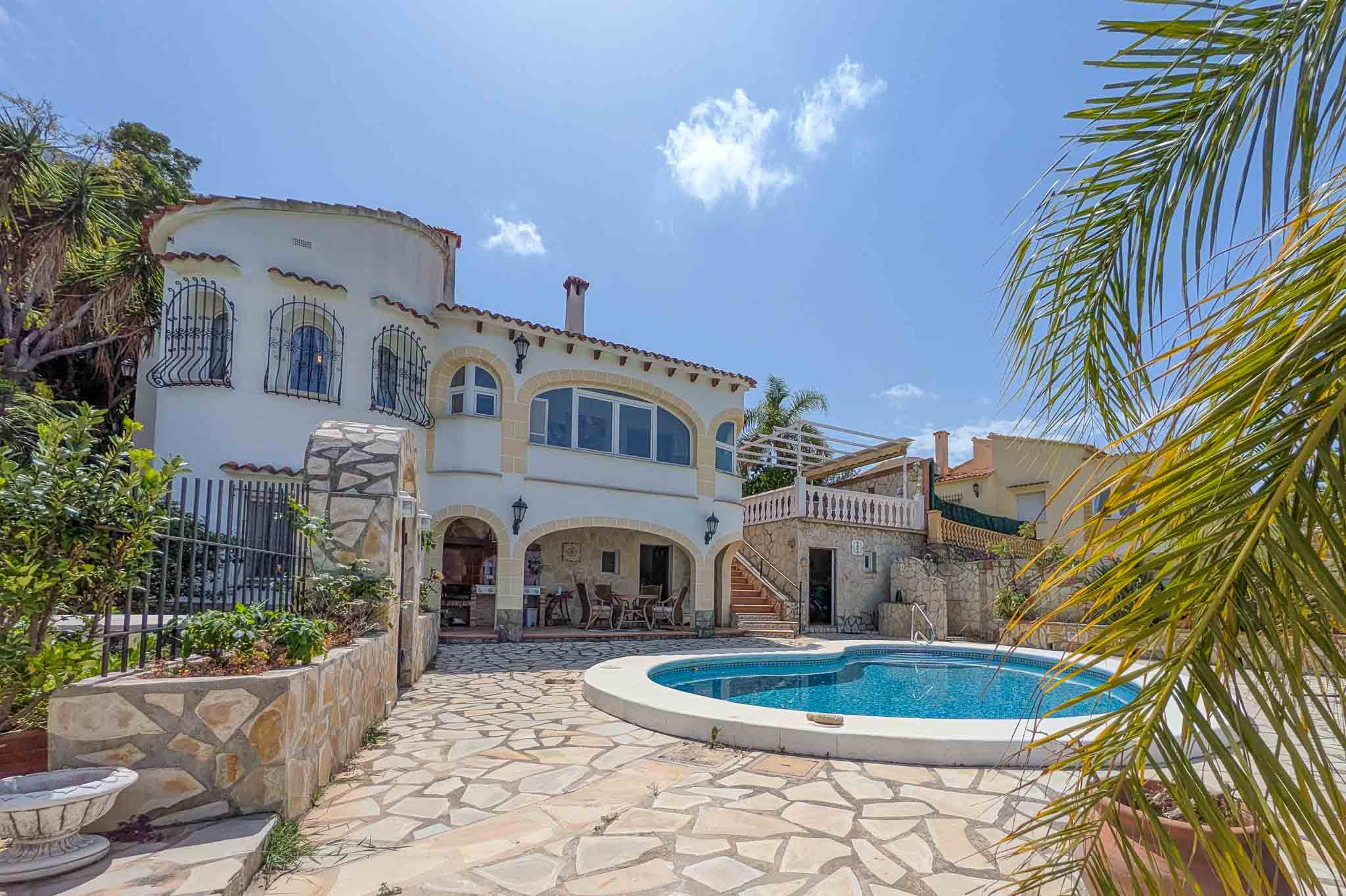 Extérieur de la villa avec piscine incurvée, terrasse en pierre naturelle et porche couvert avec cuisine d'été. La façade blanche intègre des arches et une tour latérale.