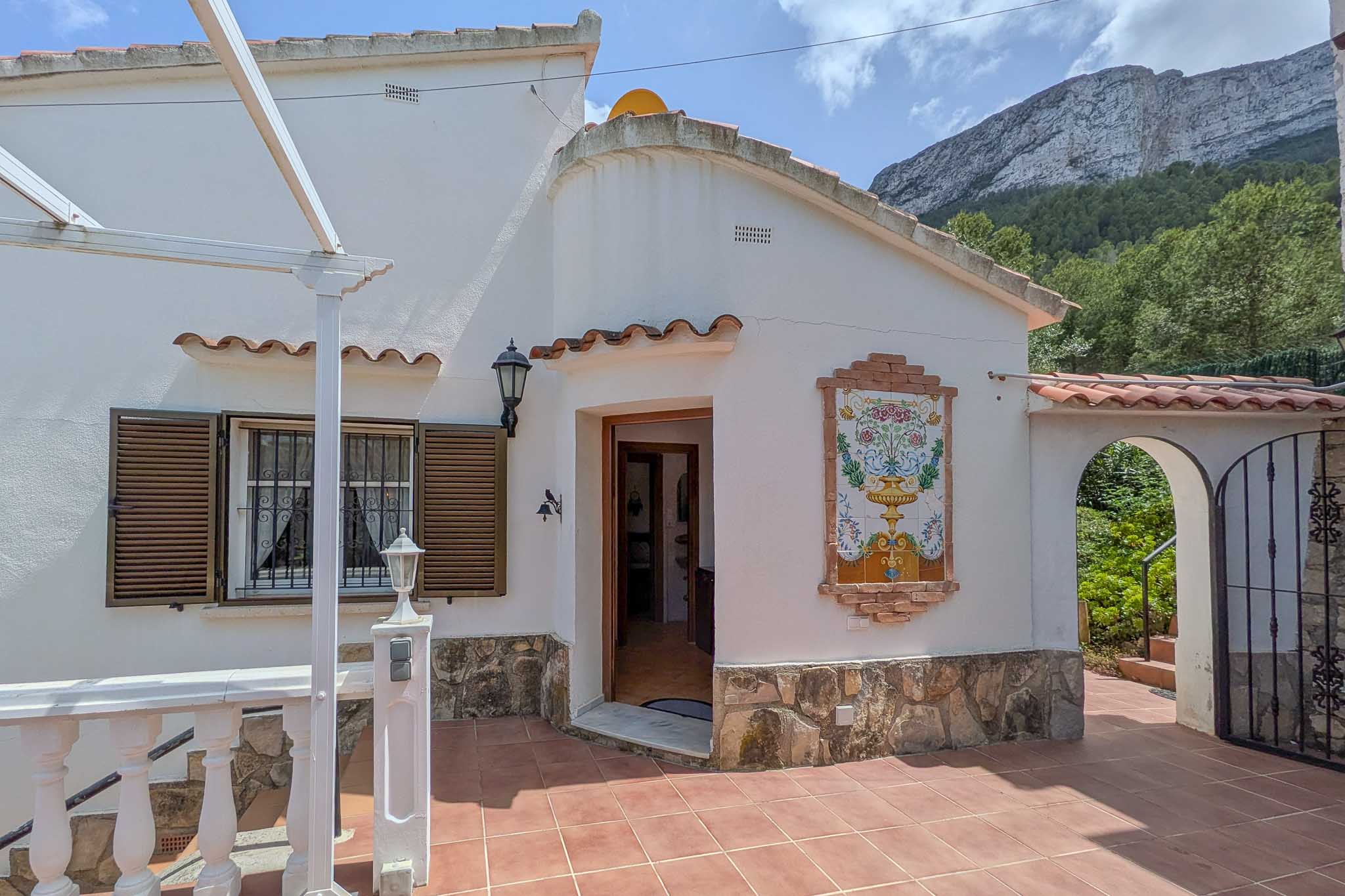 Façade blanche avec soubassement en pierre, azulejo décoratif et terrasse en terre cuite. Le mont Montgó se dessine en arrière-plan de l'entrée.