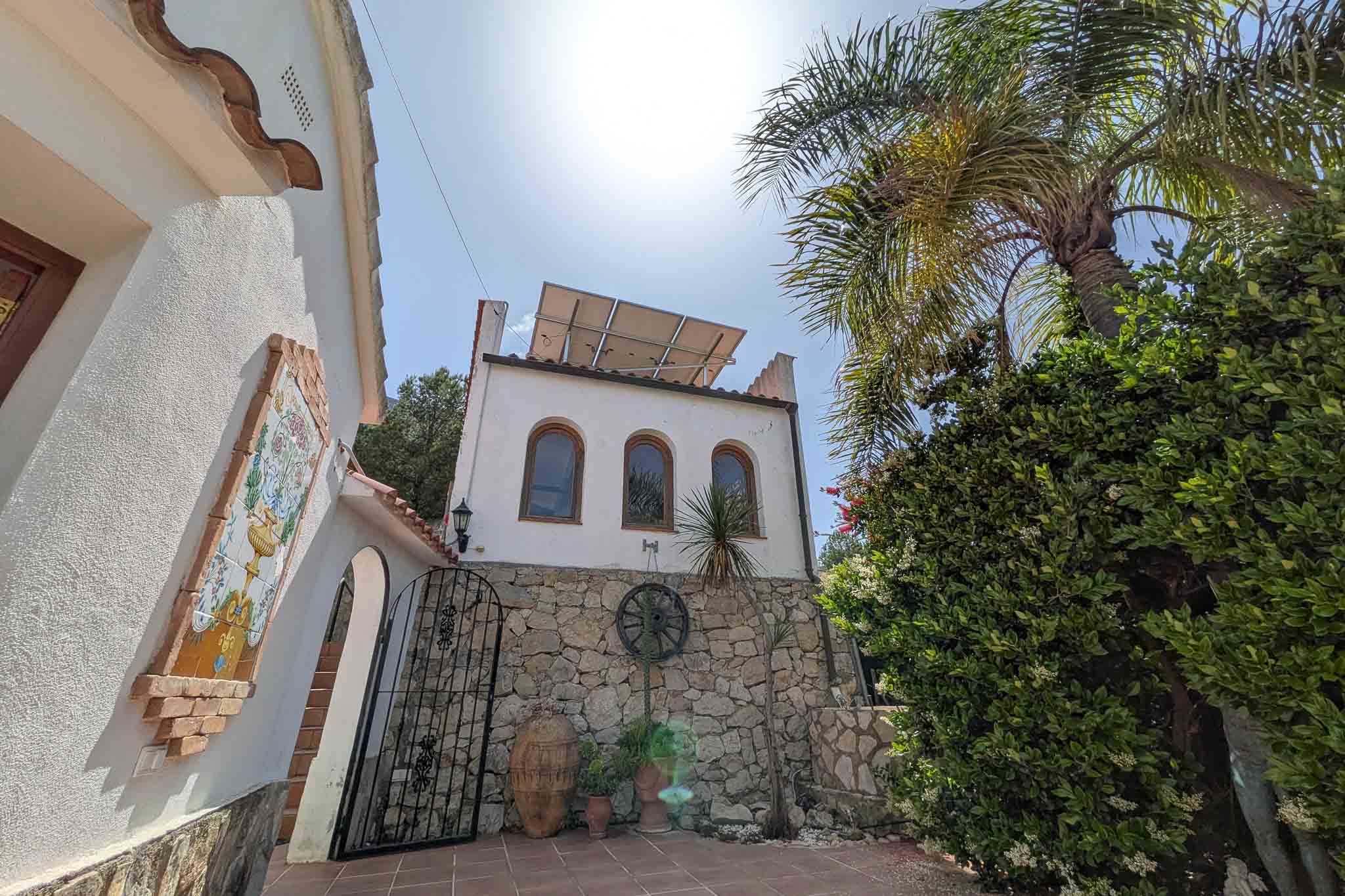 Façade méditerranéenne avec murs en pierre naturelle, panneaux solaires en toiture et détails traditionnels tels qu'une fresque en céramique et fer forgé.