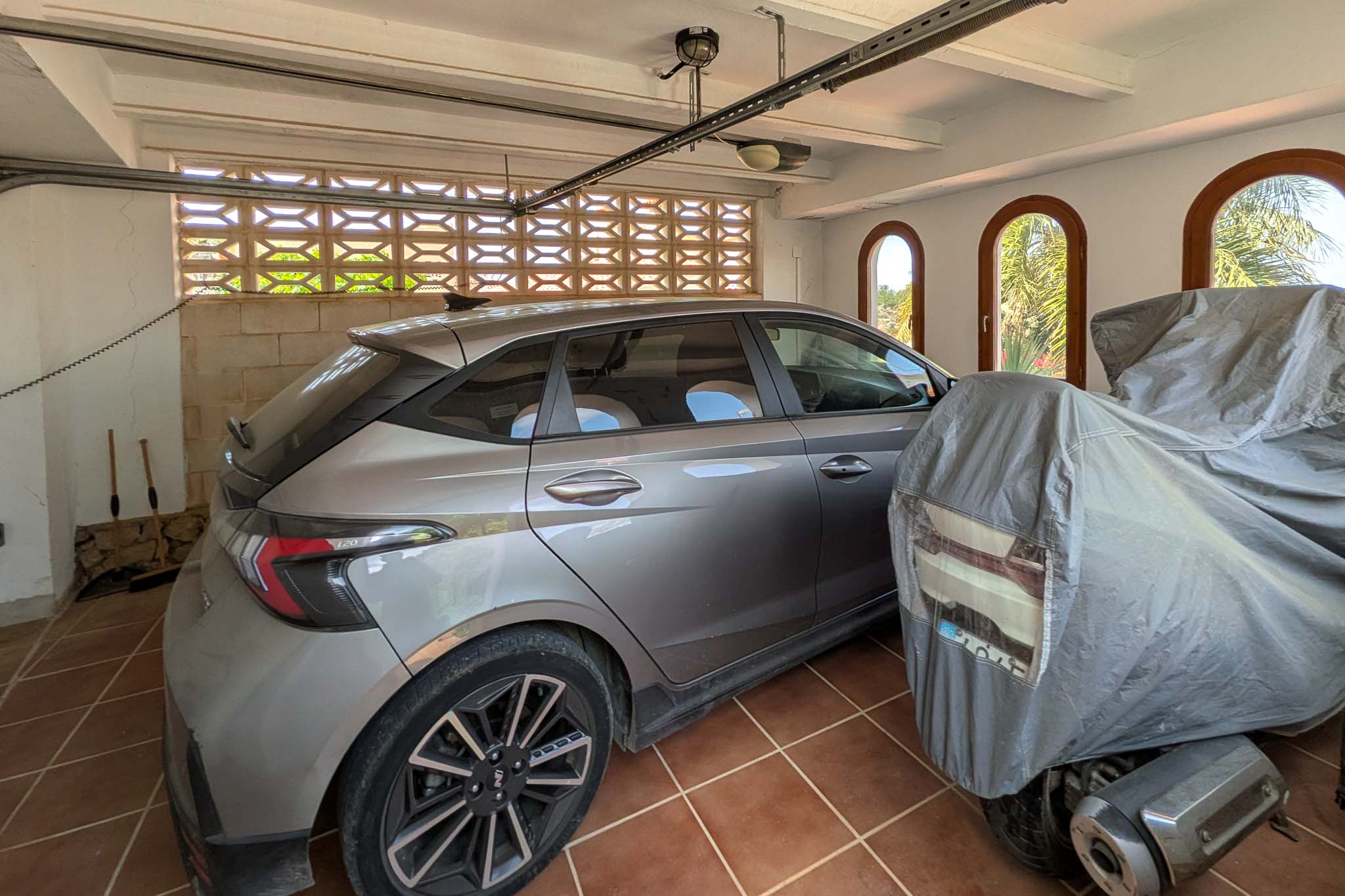 Garage couvert avec sol en grès, plafond à poutres blanches et fenêtres en arc. Un mur en treillis assure une ventilation naturelle pour l'espace de stationnement.