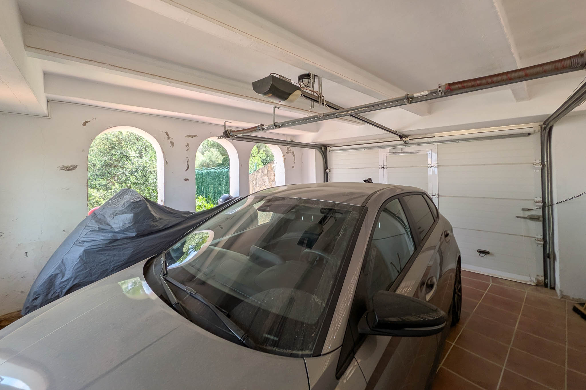 Garage fermé doté d'une porte sectionnelle automatique, d'un sol en grès et de poutres apparentes. Des arches latérales permettent une ventilation naturelle.