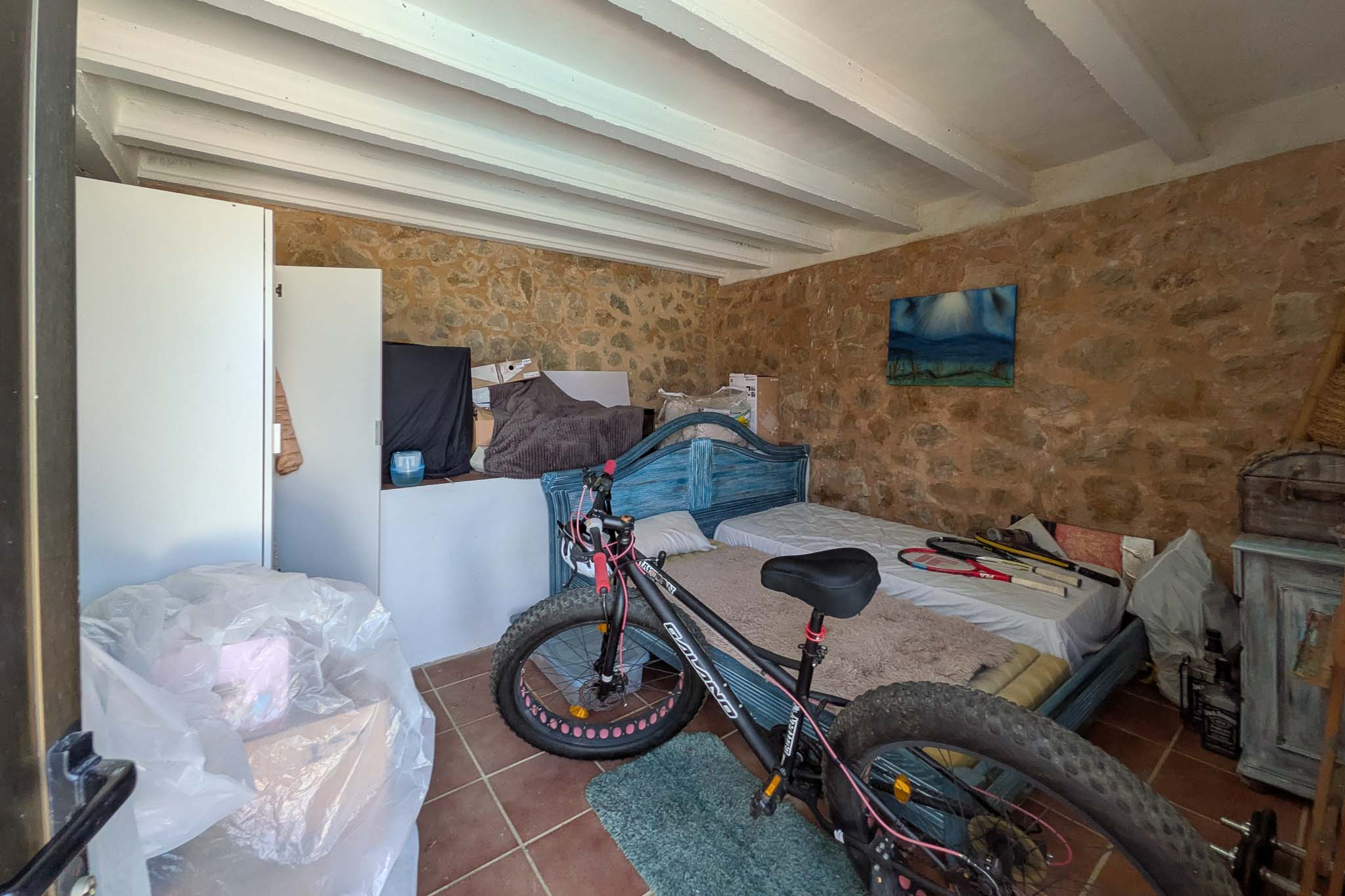 Pièce polyvalente avec murs en pierre naturelle apparente, plafonds à poutres blanches et sol en terre cuite, servant d'espace de stockage.