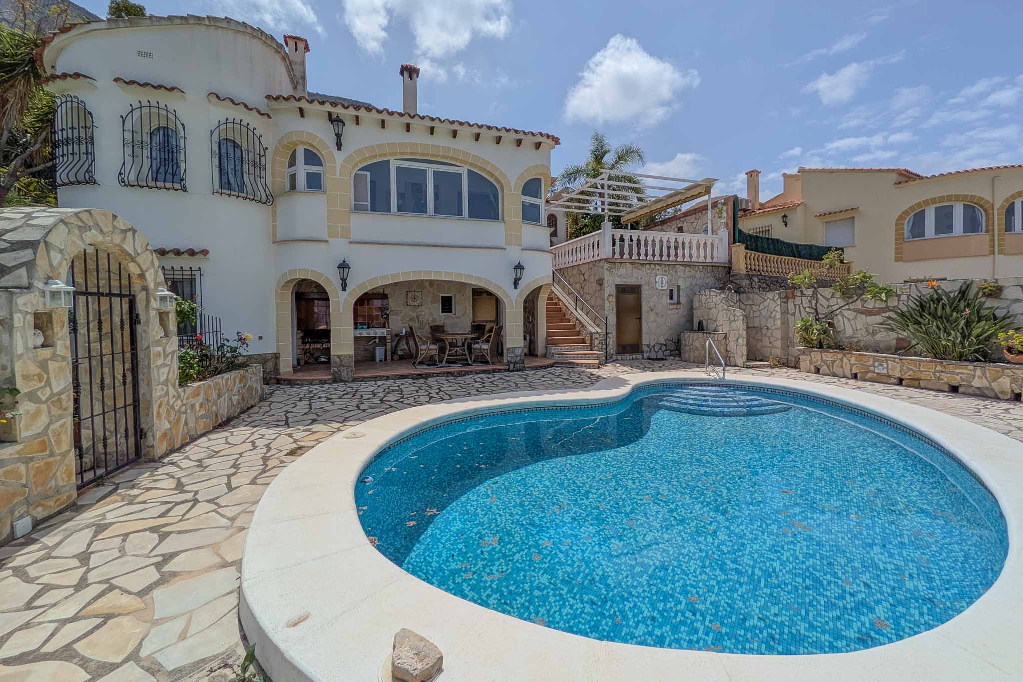 Piscine en mosaïque bleue entourée d'une terrasse en pierre, avec un porche couvert abritant une cuisine d'été et un coin repas.