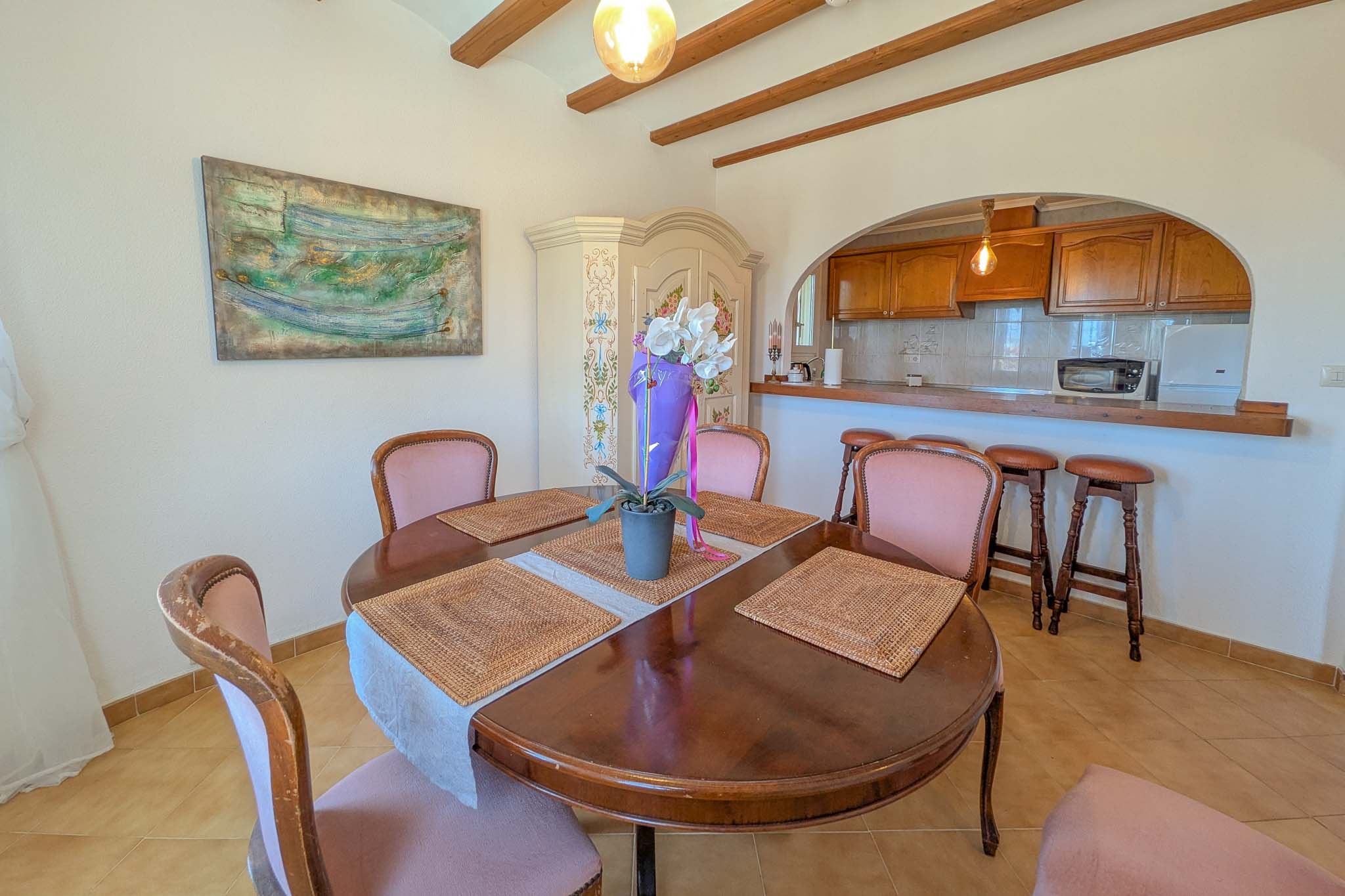 Salle à manger avec table ronde en bois et poutres apparentes, ouverte sur une cuisine via une arche avec bar et tabourets de style rustique.