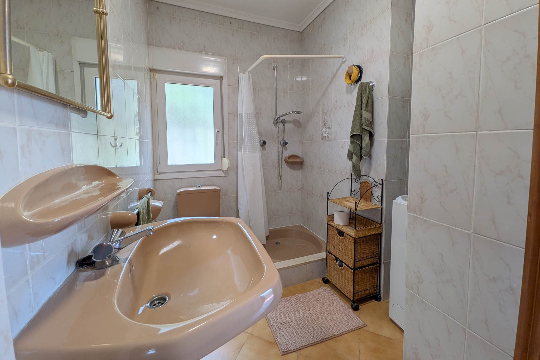 Salle de bain fonctionnelle avec sanitaires de couleur saumon, receveur de douche avec rideau, murs carrelés et fenêtre pour ventilation naturelle.