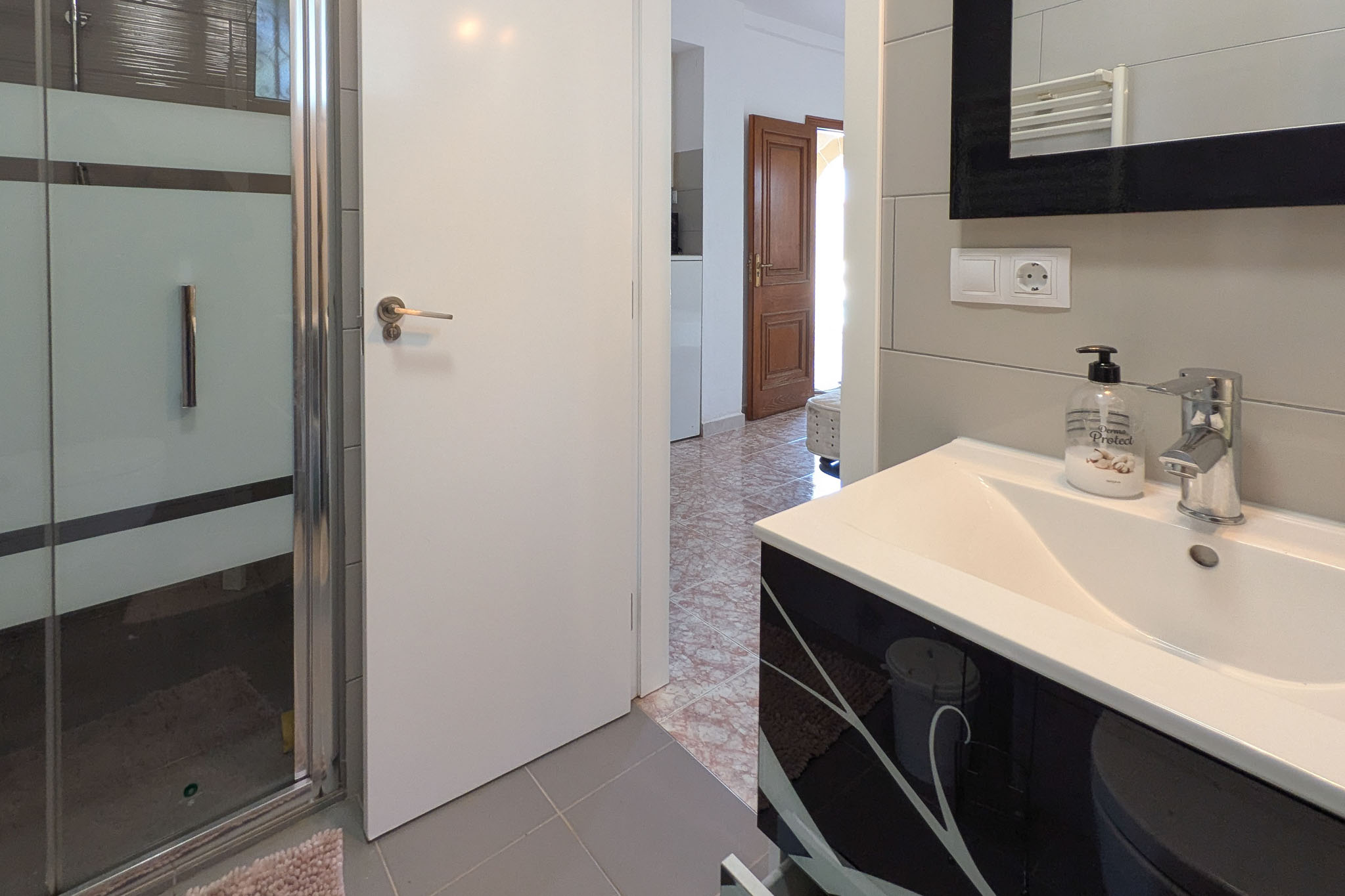 Salle de bain moderne avec meuble vasque noir à motifs, cabine de douche vitrée et carrelage gris. La porte blanche s'ouvre sur un couloir au sol en terre cuite.