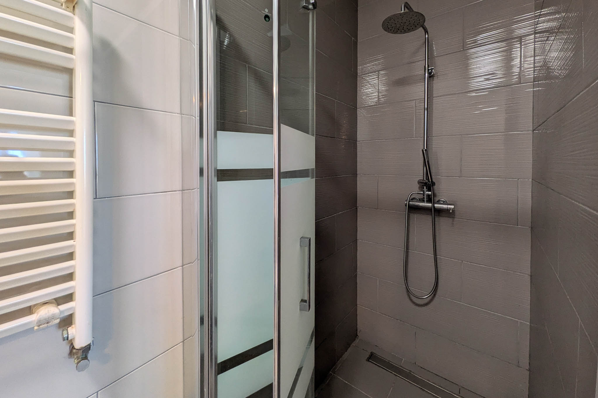 Salle d'eau avec douche à l'italienne, paroi vitrée sérigraphiée, robinetterie chromée et radiateur sèche-serviettes blanc sur carrelage contrasté.