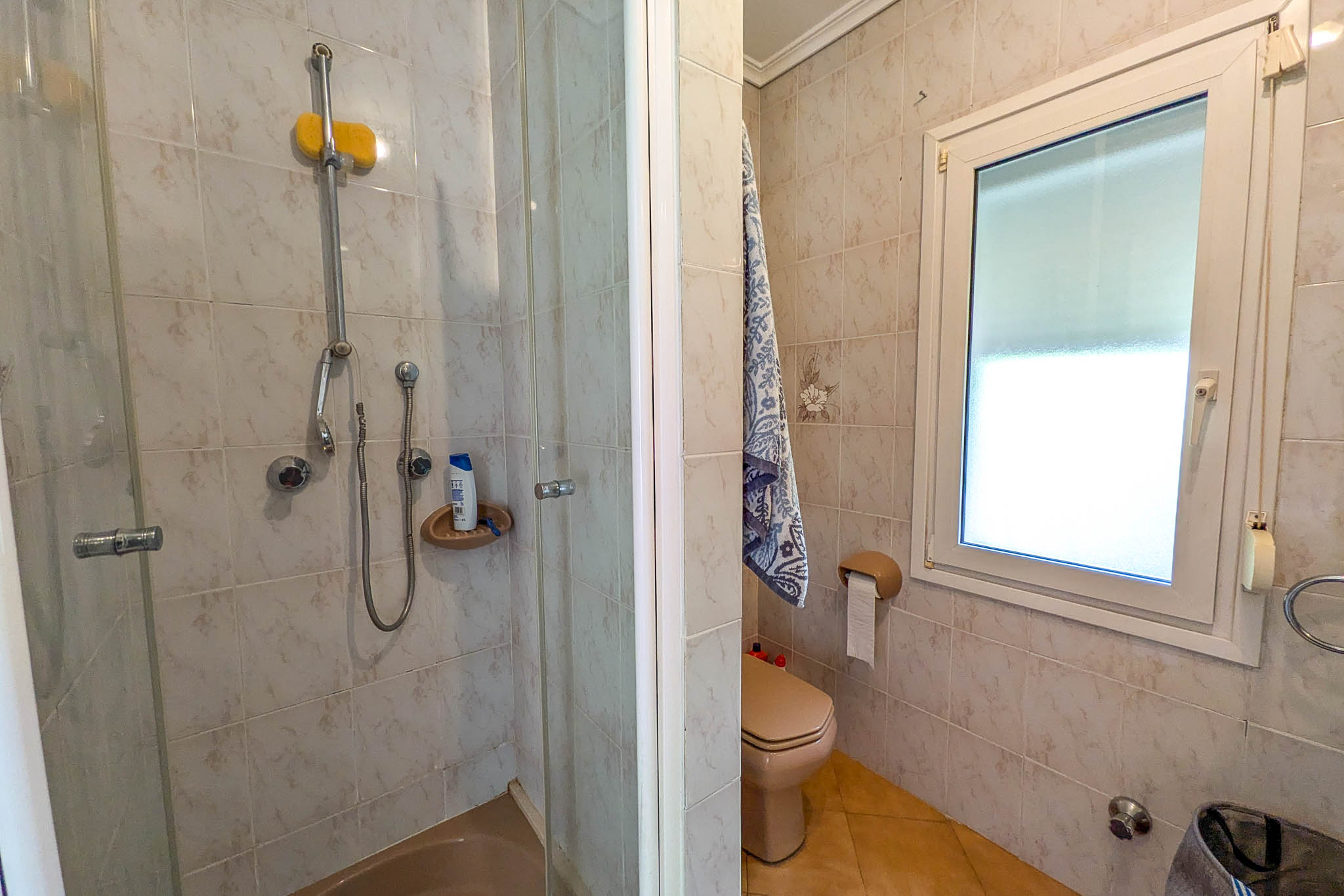 Salle d'eau comprenant une douche à paroi vitrée, des toilettes de couleur crème et une fenêtre en PVC blanc. Les murs sont revêtus de carreaux de céramique à motifs discrets.