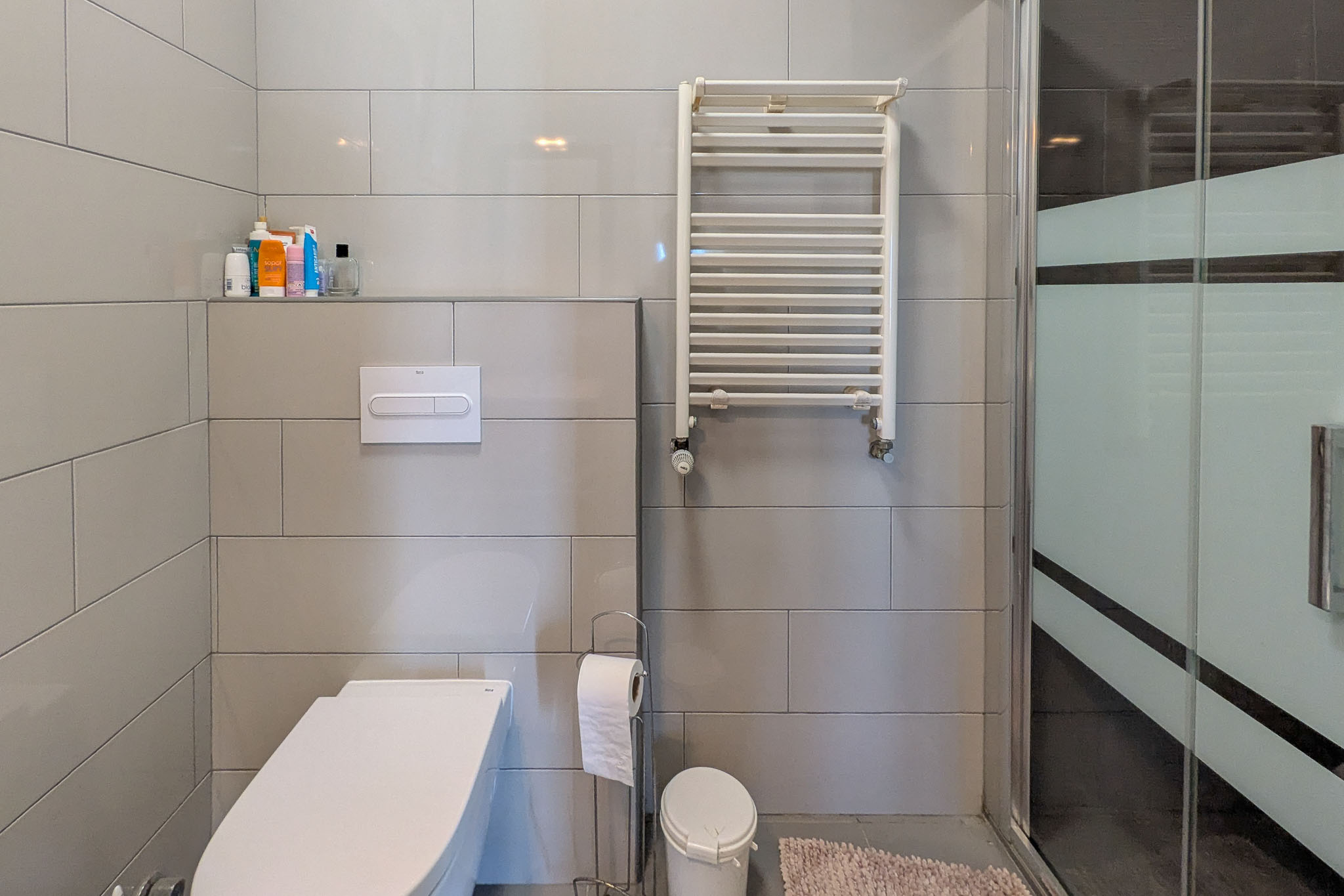 Salle d'eau moderne avec WC suspendu, carrelage mural gris, radiateur sèche-serviettes blanc et paroi de douche en verre avec bande dépolie.