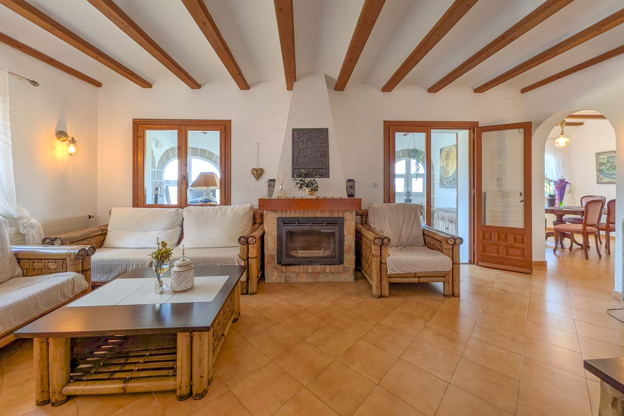 Salon rustique avec cheminée en pierre, poutres en bois apparentes et sol en terre cuite. Le mobilier en bambou et l'arche vers la salle à manger créent une ambiance méditerranéenne.