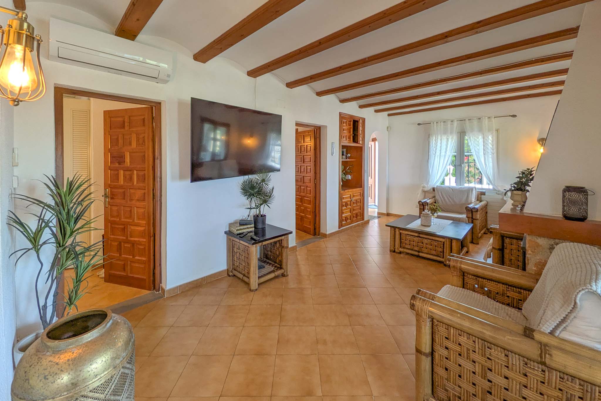 Salon rustique avec poutres apparentes, sol en terre cuite et mobilier en bambou. Comprend la climatisation et des portes en bois sculpté menant aux chambres.