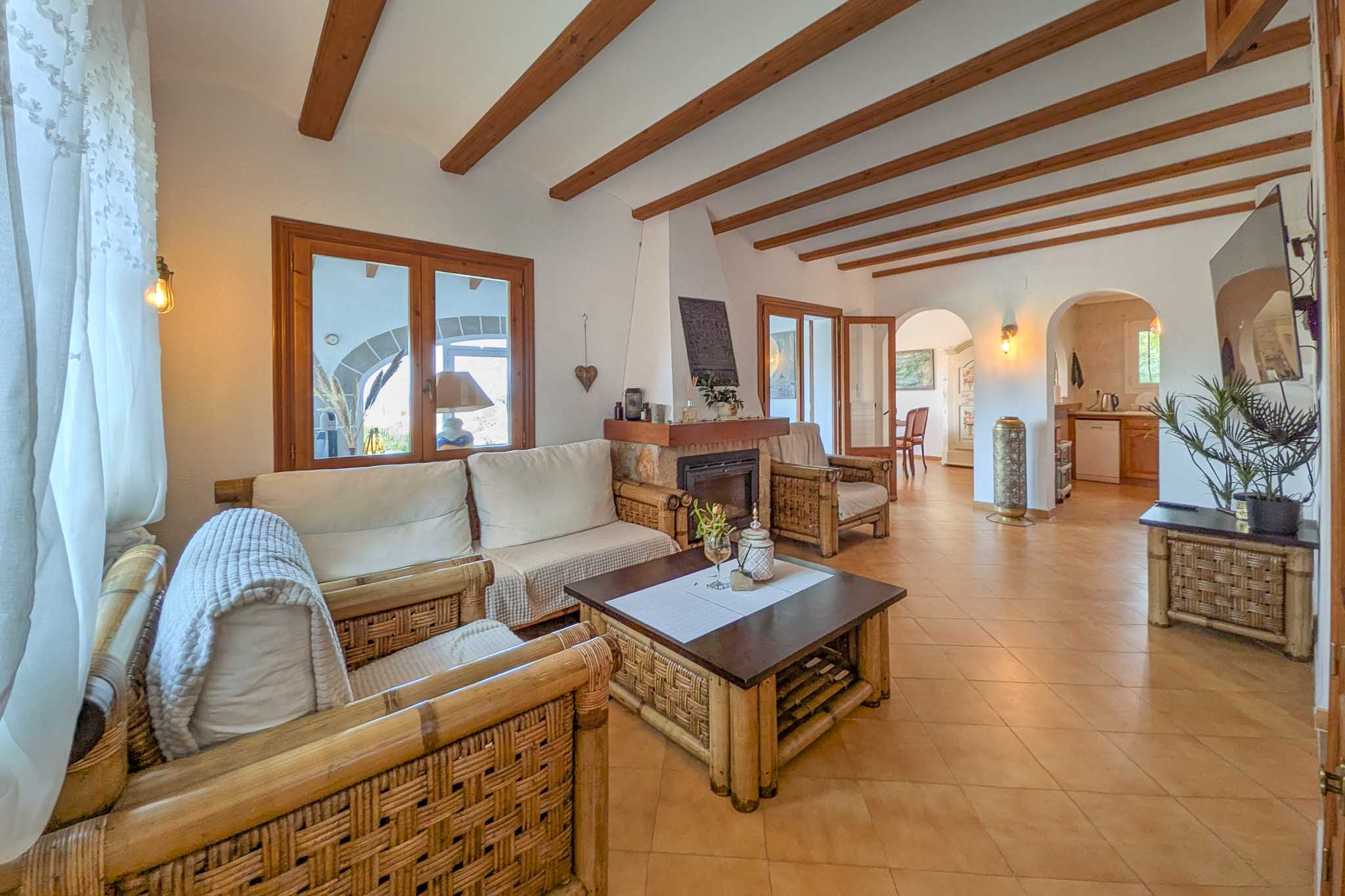 Salon rustique avec poutres en bois apparentes, cheminée en pierre et mobilier en bambou. L'espace communique par des arches avec la salle à manger et la cuisine.