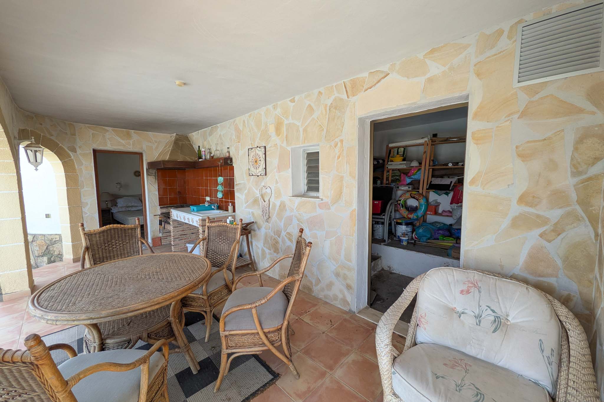Terrasse couverte au rez-de-chaussée avec murs en pierre naturelle, cuisine d'été intégrée et coin repas en osier à côté de l'espace de rangement.