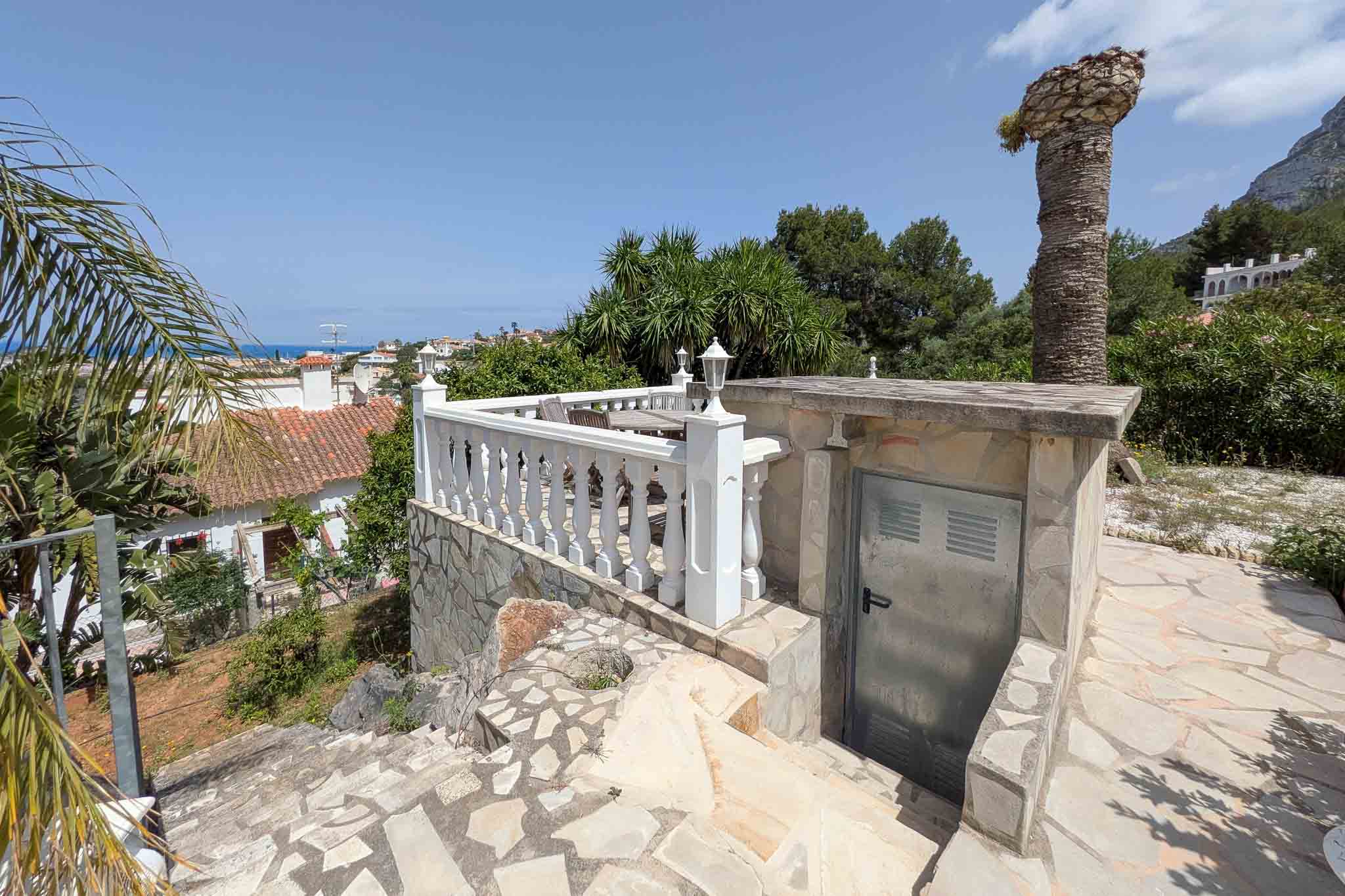 Terrasse extérieure avec balustrades blanches et sol en pierre naturelle, offrant une vue dégagée sur la mer et un accès à un local technique.