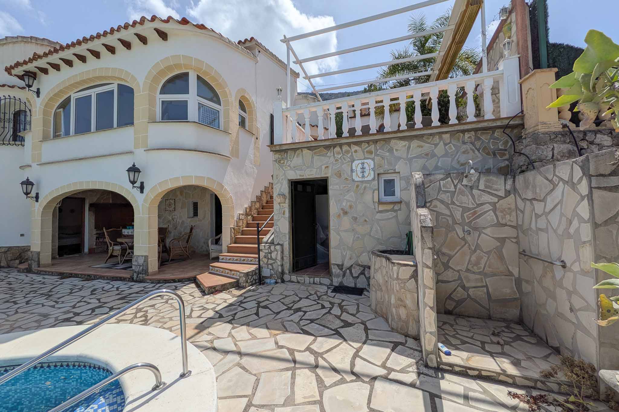 Terrasse extérieure en pierre naturelle avec douche intégrée, accès à la piscine et escalier menant au niveau supérieur avec balustrade blanche.