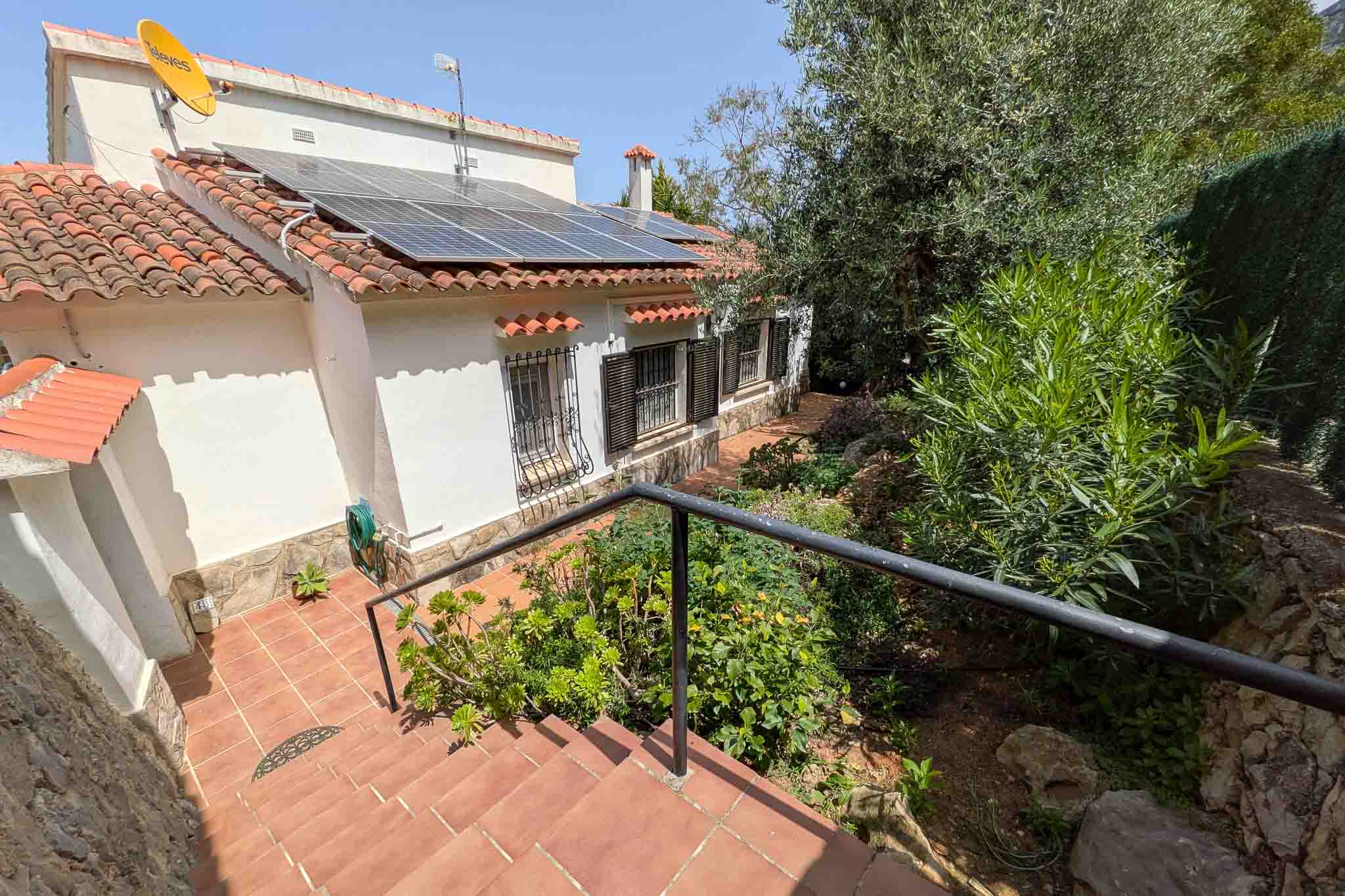 Vue extérieure de la façade blanche avec panneaux photovoltaïques sur le toit, escalier en terre cuite et jardin méditerranéen luxuriant.