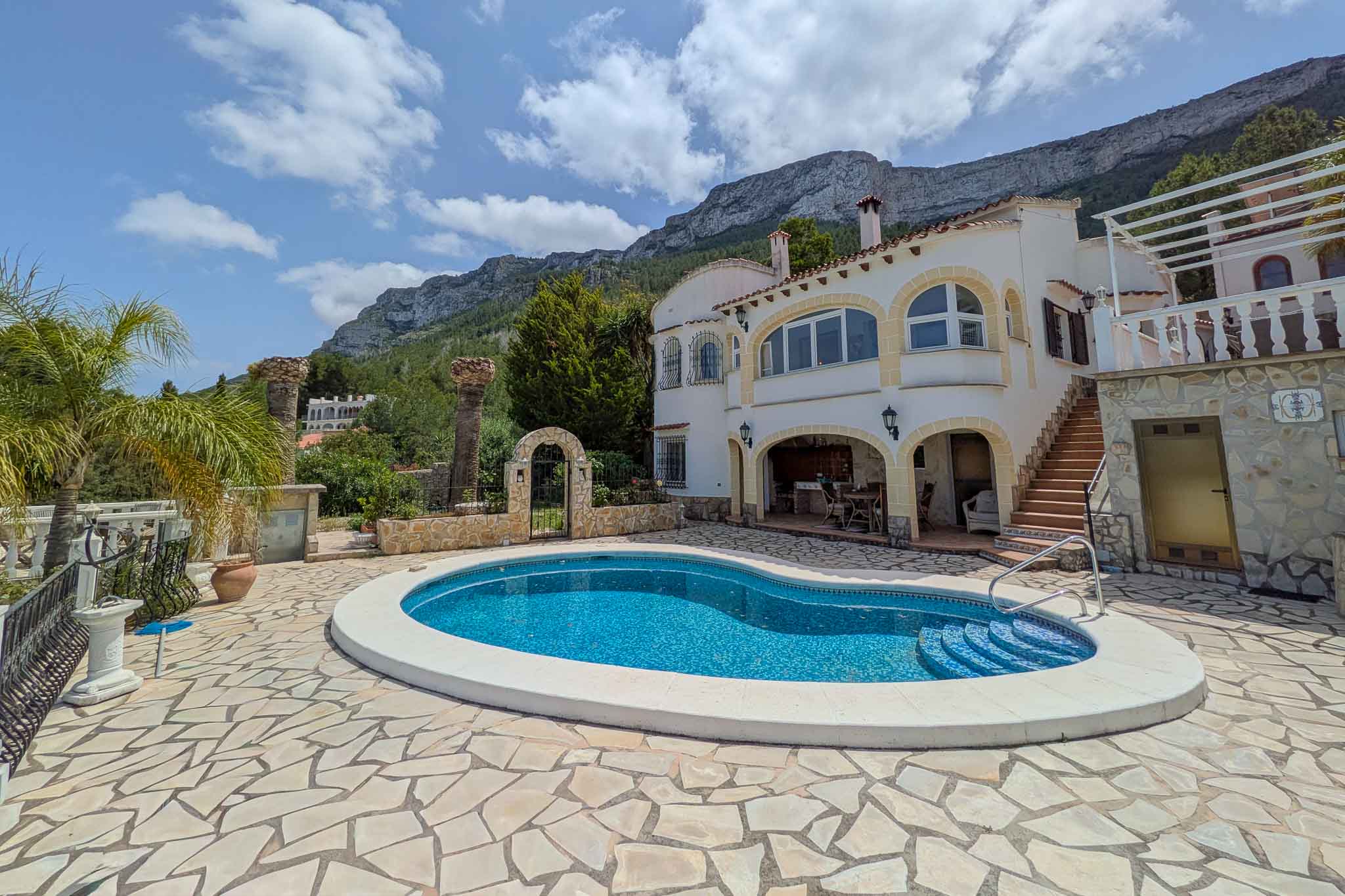 Vue extérieure de la piscine de forme libre avec terrasse en pierre naturelle, façade blanche aux arcs décoratifs et le mont Montgó en arrière-plan.