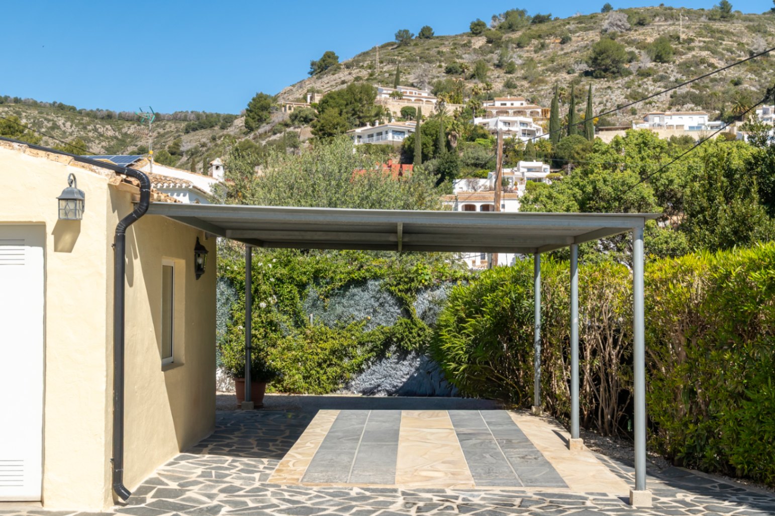 Carport métallique avec toit plat et sol en pierre naturelle, situé à côté de la villa avec vue sur les collines verdoyantes de Jávea.