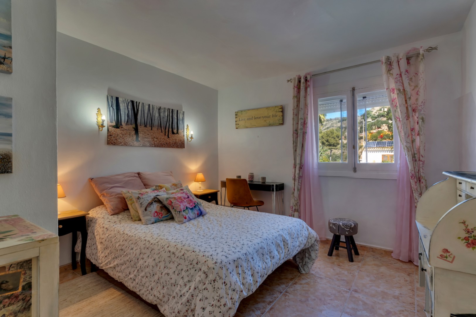 Chambre avec sol en céramique, coin bureau et fenêtre vue sur l'extérieur. Elle présente une décoration classique avec des textiles floraux et un bureau secrétaire.