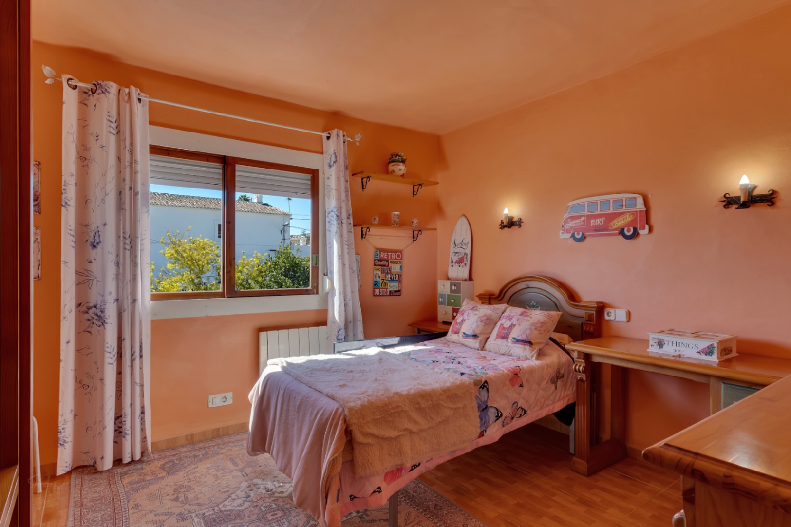 Chambre lumineuse avec murs saumon, mobilier en bois massif, bureau intégré et fenêtre. Équipée d'un radiateur électrique et d'un sol en carrelage.