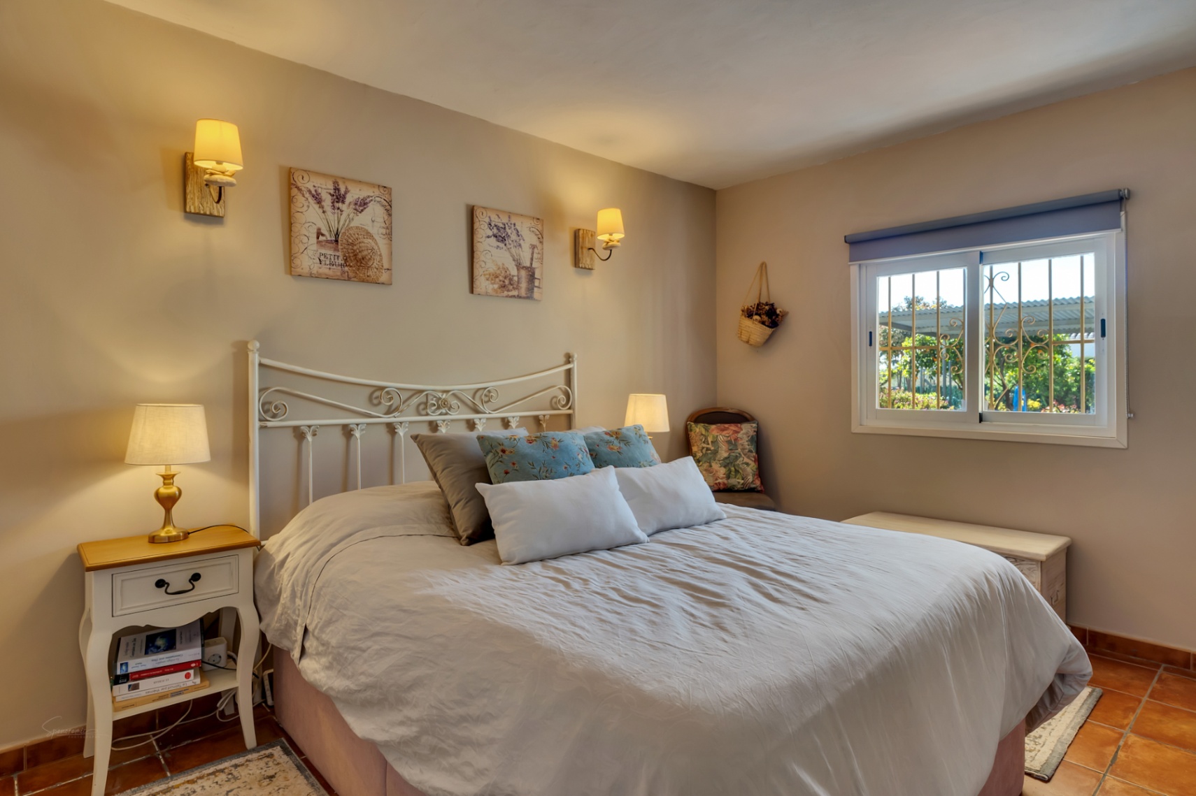 Chambre lumineuse avec tête de lit en fer forgé blanc, sol en terre cuite et appliques murales. Une fenêtre avec grilles de sécurité apporte une lumière naturelle.
