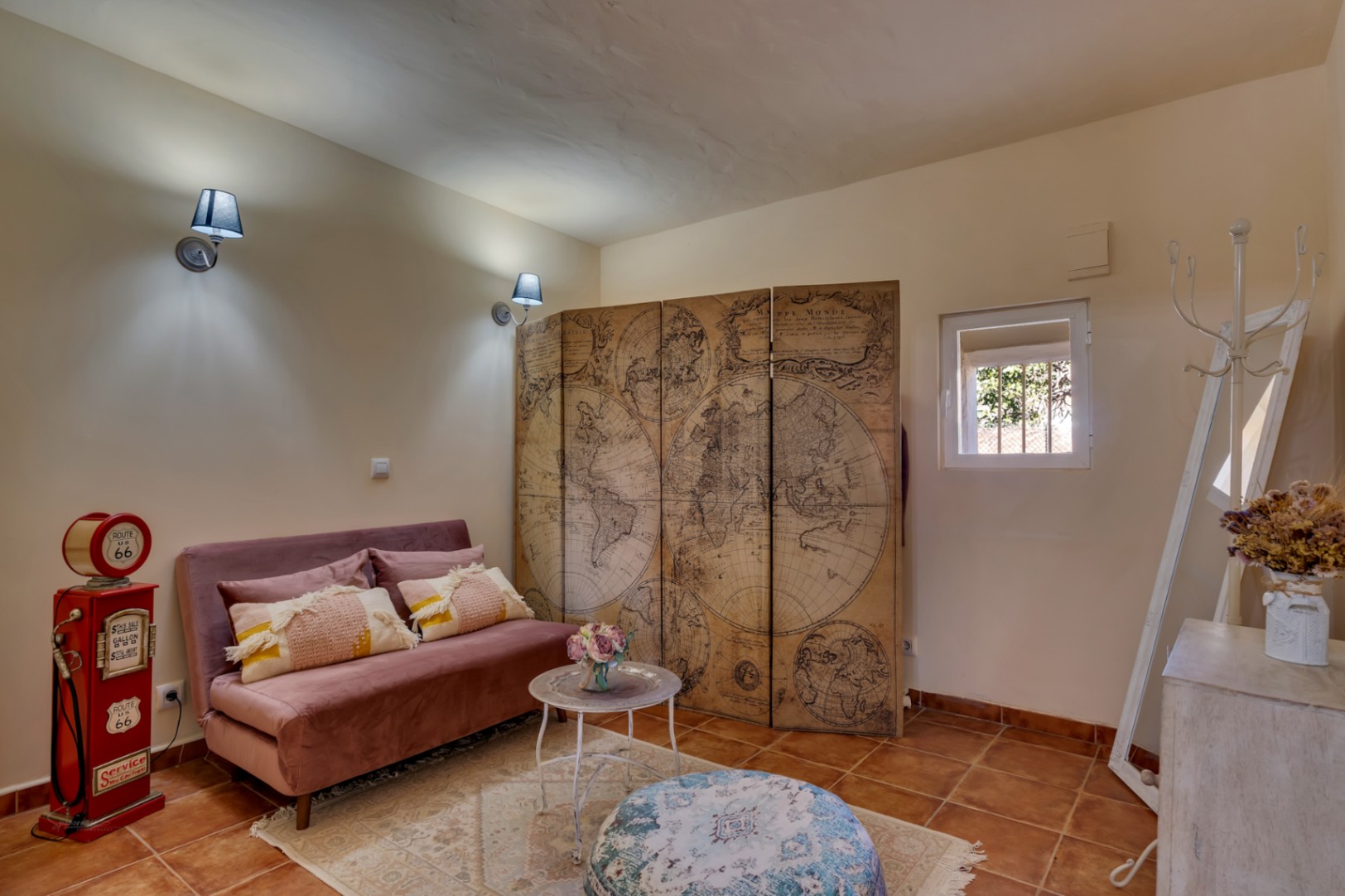 Coin salon avec sol en terre cuite, canapé en velours rose, paravent décoratif représentant une carte du monde ancienne et appliques murales classiques.