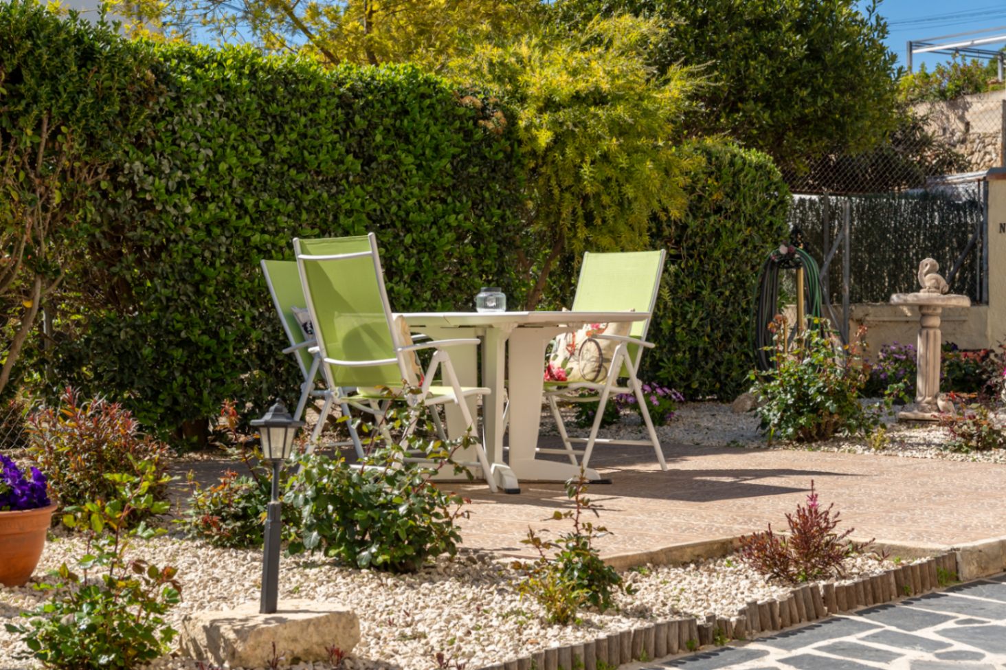 Coin salon extérieur avec table blanche et chaises vertes sur terrasse en pierre, entouré de haies denses et d'un jardin méditerranéen avec gravier et fleurs.