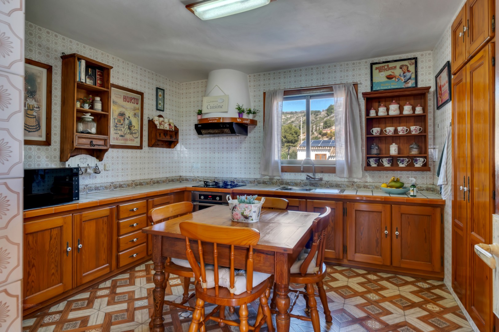 Cuisine rustique avec meubles en bois, table à manger centrale et murs carrelés. Une fenêtre offre une vue sur les collines environnantes et apporte de la lumière naturelle.