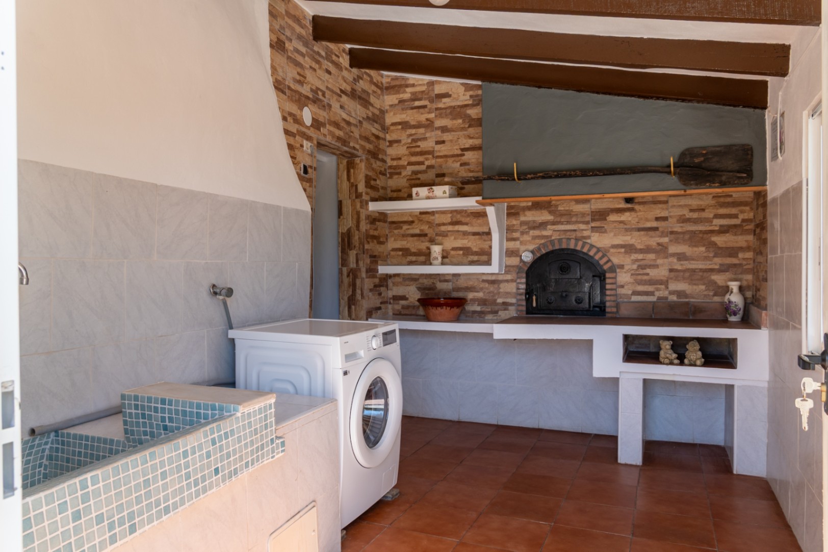 Espace buanderie et cuisine rustique avec four à bois traditionnel, évier en mosaïque bleue, lave-linge et plafonds à poutres apparentes.