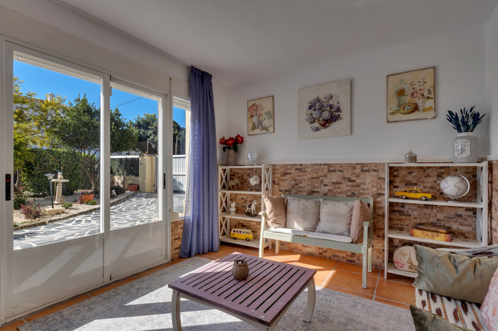 Espace de vie lumineux avec sol en terre cuite, soubassement en pierre naturelle et baies vitrées offrant un accès direct au jardin pavé.
