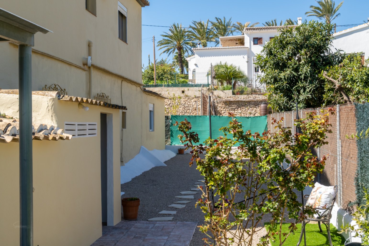 Espace extérieur latéral avec chemin en gravier, dalles de passage, végétation locale et une petite construction annexe fonctionnelle.