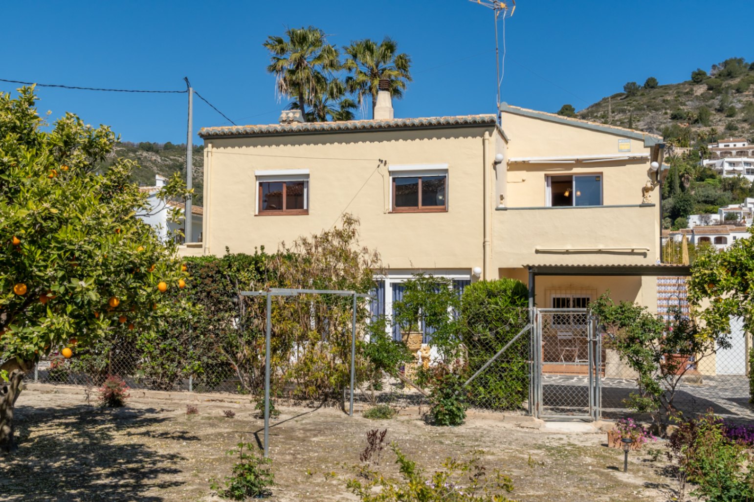 Façade extérieure de la villa aux tons crème, entourée d'un jardin avec des orangers et des palmiers, située au pied d'une colline.