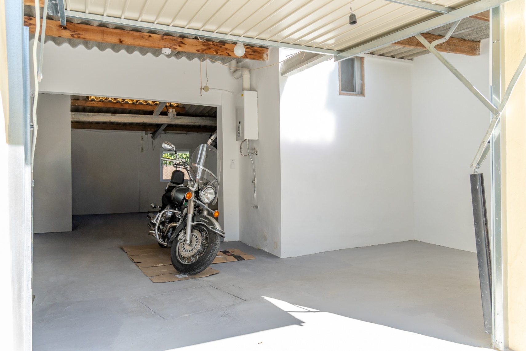 Garage fermé avec sol en béton, murs blancs et poutres en bois apparentes. L'espace comprend un chauffe-eau mural et offre une grande capacité de rangement pour véhicules.