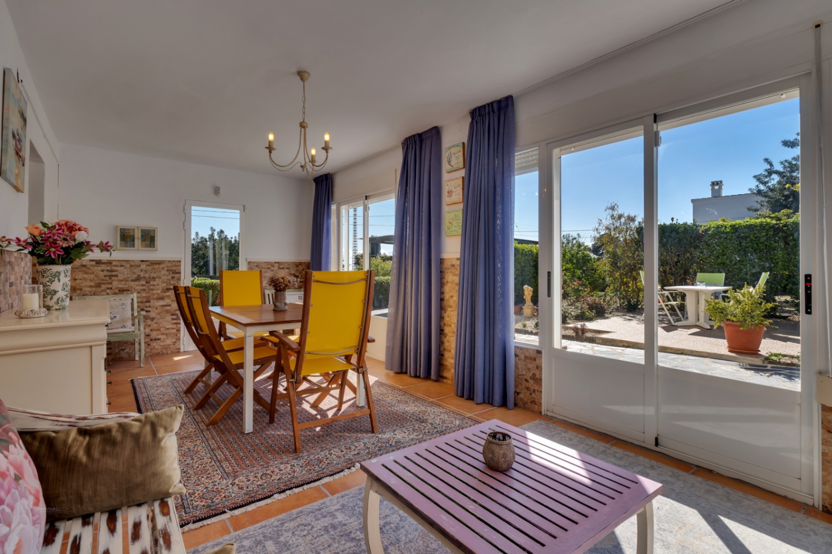 Salle à manger lumineuse avec chaises jaunes, murs en pierre partielle et grandes baies vitrées coulissantes offrant un accès direct au jardin méditerranéen.