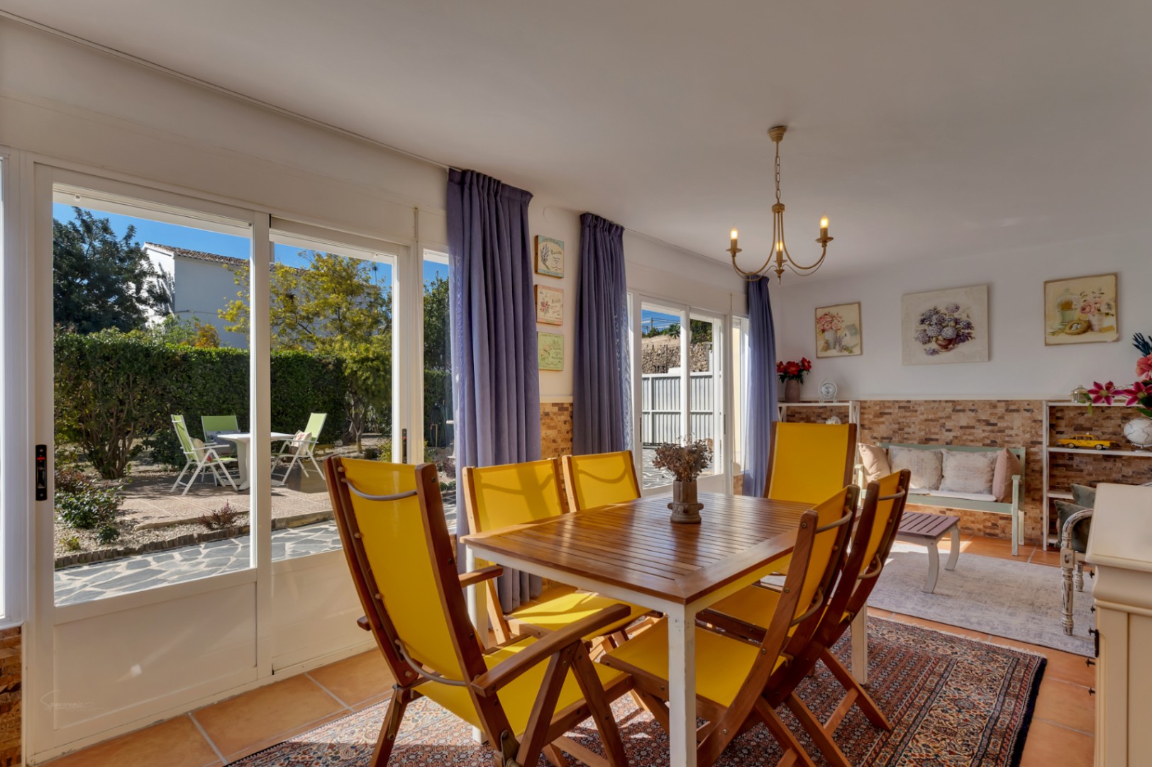 Salle à manger lumineuse avec table en bois, chaises jaunes et accès direct au jardin par de grandes baies vitrées sur un sol en terre cuite.