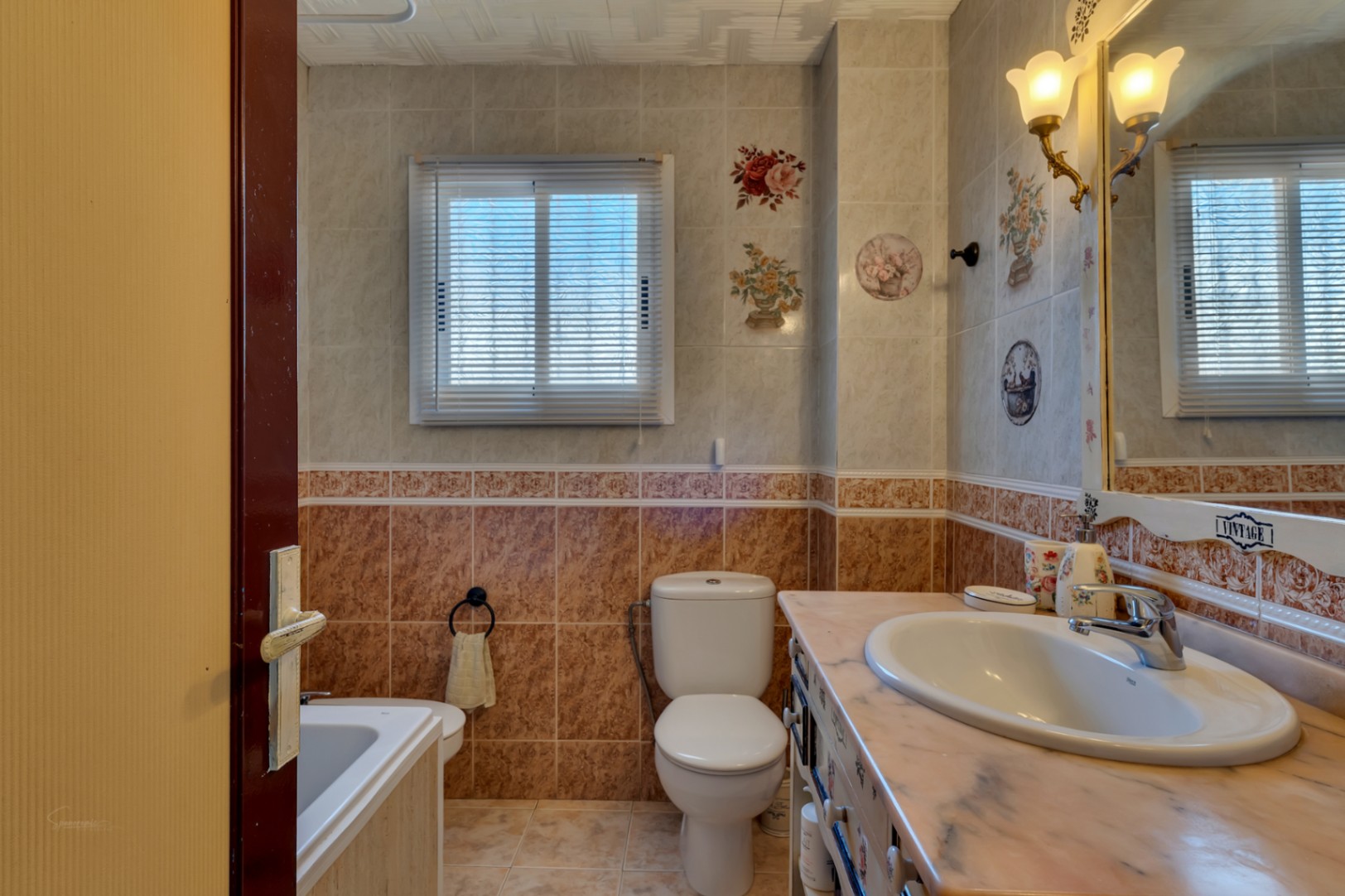 Salle de bain complète avec carrelage décoratif, plan de toilette en marbre, appliques murales classiques et fenêtre avec stores intégrés.