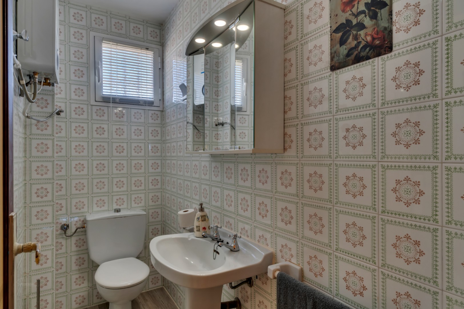 Salle de bain fonctionnelle avec murs carrelés à motifs, lavabo sur colonne, toilettes et armoire de toilette à miroir avec éclairage intégré.