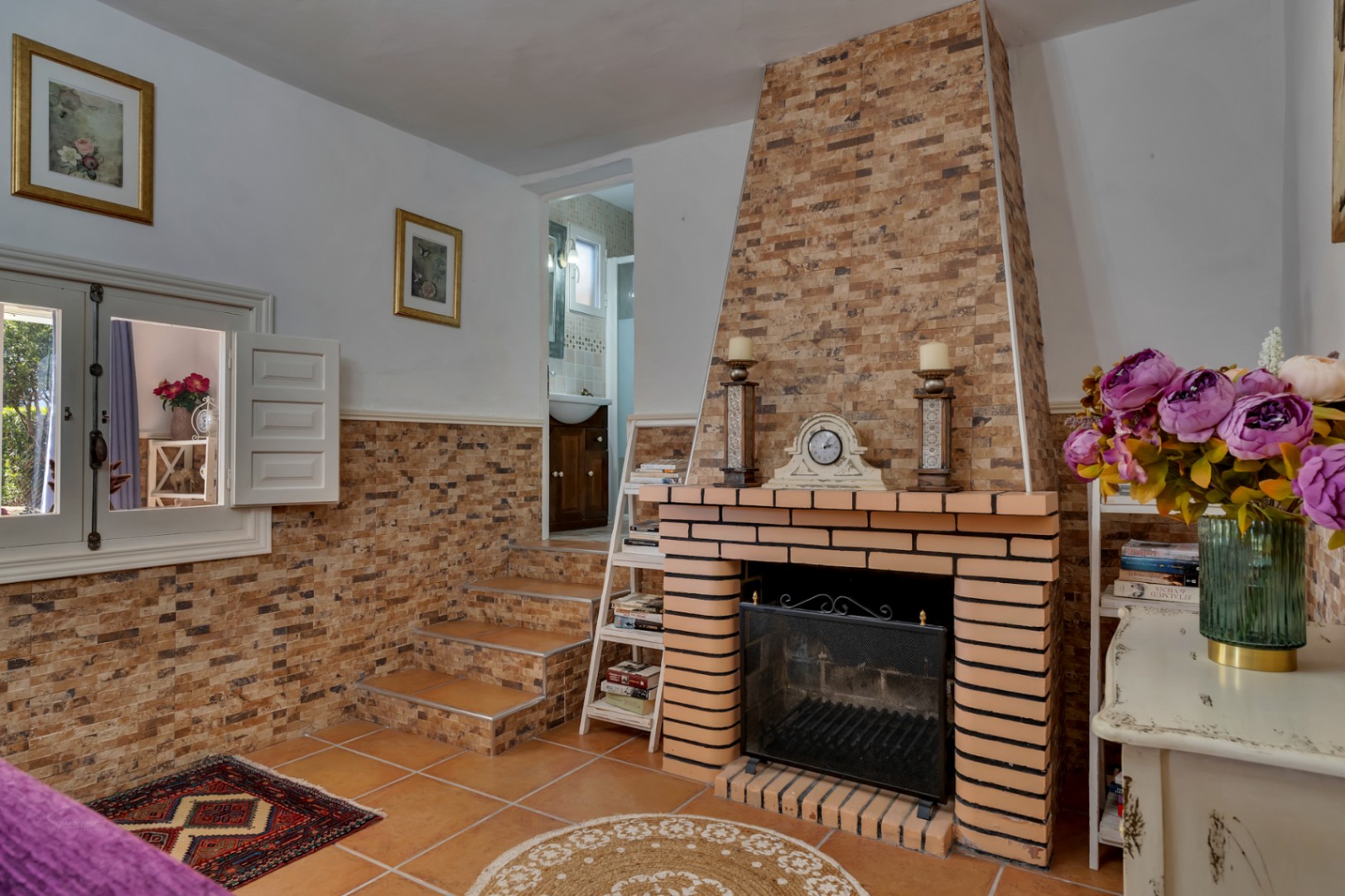 Salon avec cheminée en briques et parement pierre, sols en terre cuite et marches menant à une salle de bain. Des volets intérieurs blancs complètent ce décor méditerranéen.