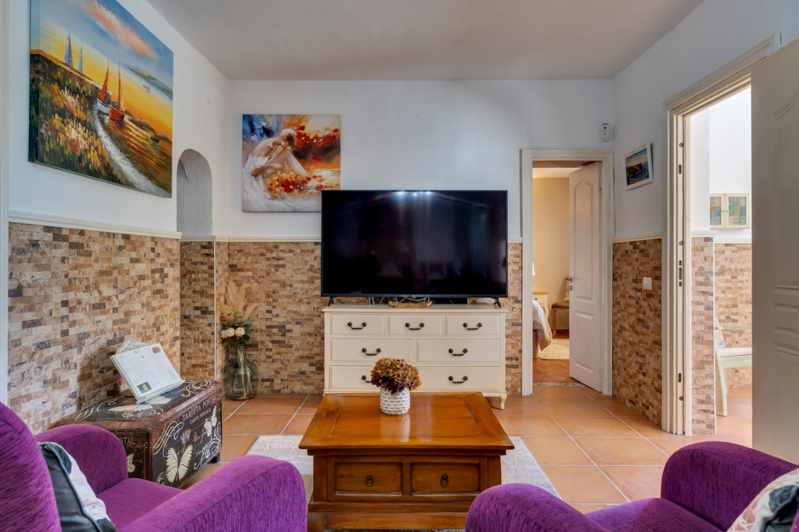 Salon avec fauteuils violets, soubassement en pierre naturelle, sol en terre cuite et meuble TV blanc ouvrant sur une chambre.