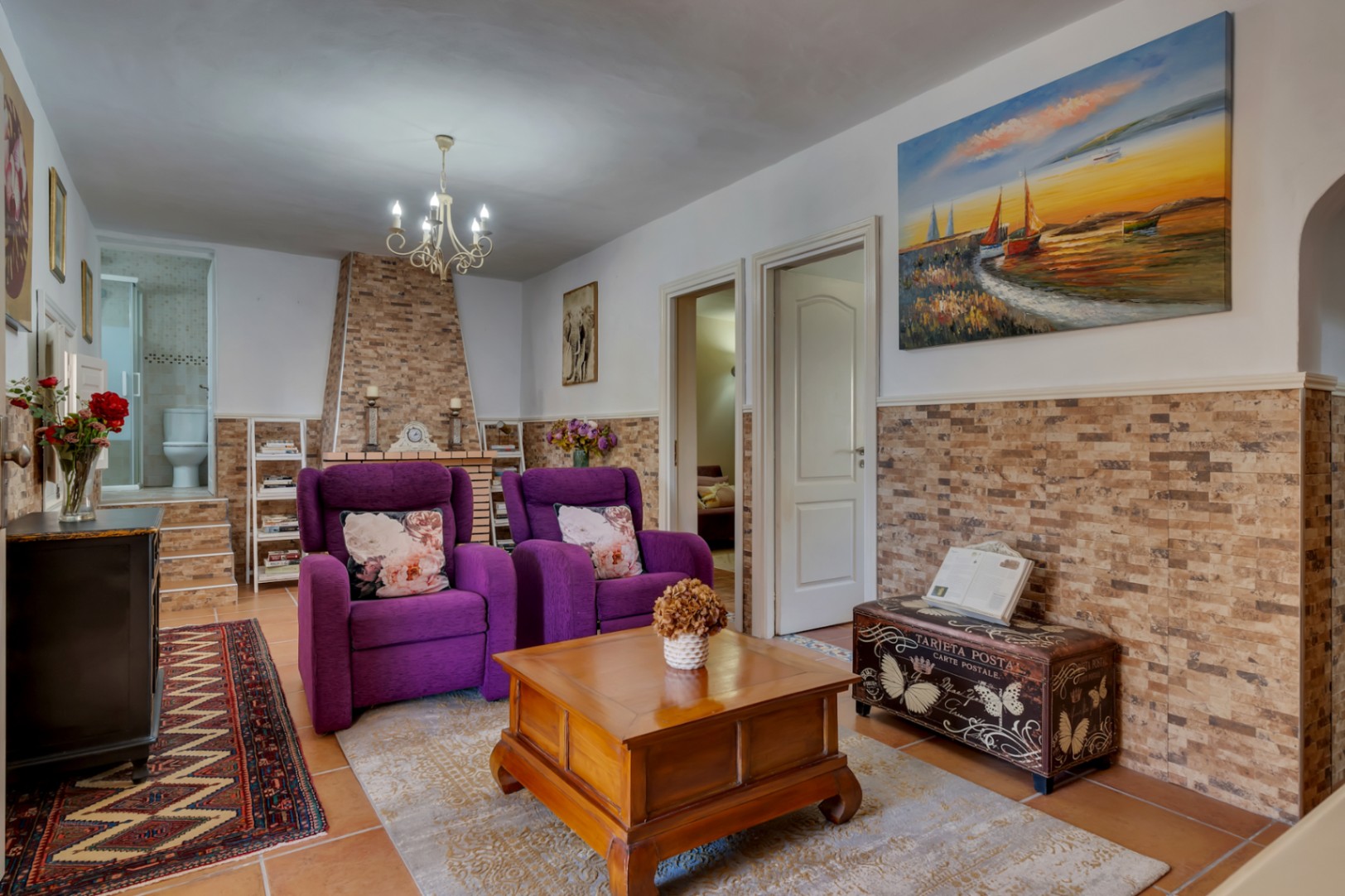 Salon chaleureux avec cheminée en pierre, fauteuils violets et sol en terre cuite, offrant un accès direct à une salle de bain attenante.