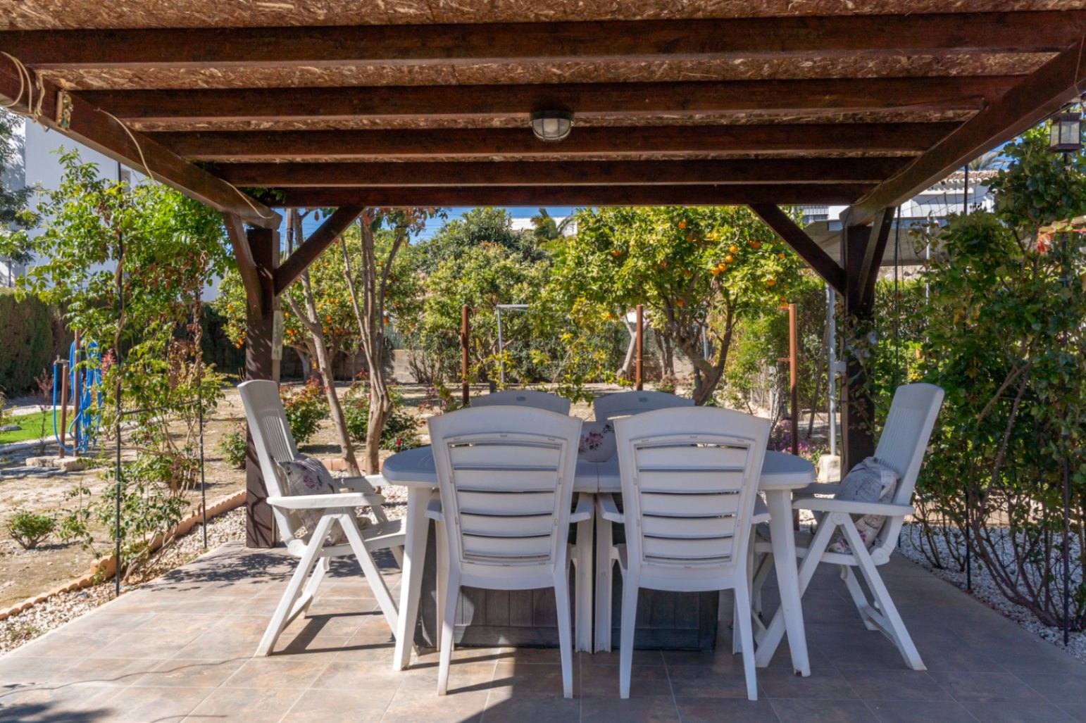 Terrasse extérieure couverte par une pergola en bois, avec coin repas et vue sur le jardin méditerranéen planté d'agrumes.