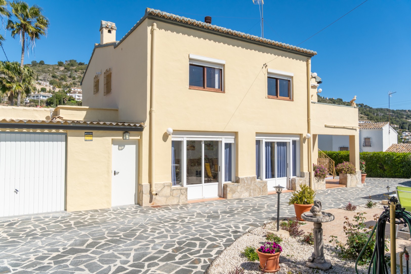 Vue extérieure de la villa avec façade crème, soubassement en pierre et grand patio pavé. Comprend un garage fermé et de larges menuiseries blanches au rez-de-chaussée.