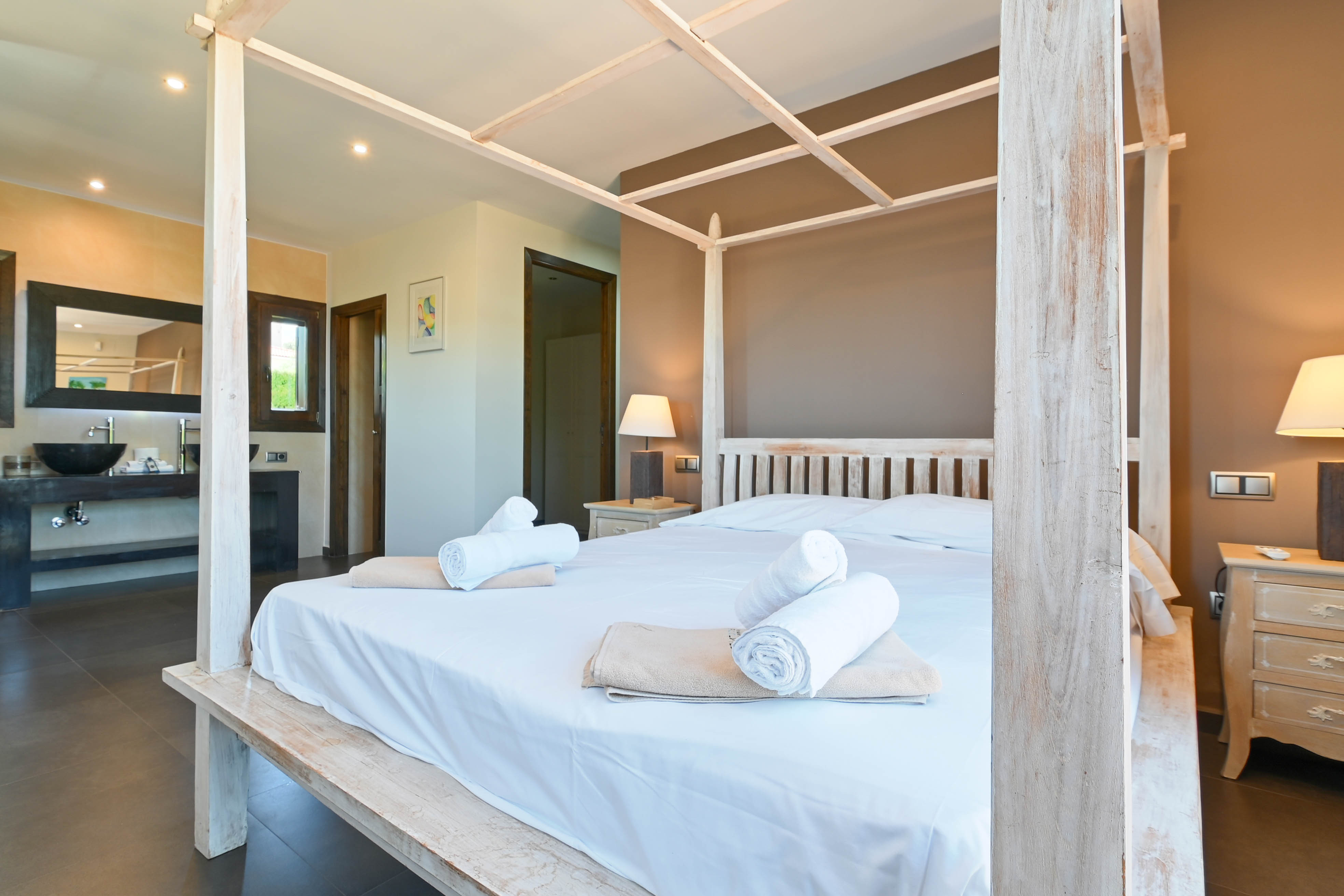 Chambre avec lit à baldaquin en bois blanc, serviettes roulées et salle de bain avec lavabo moderne.