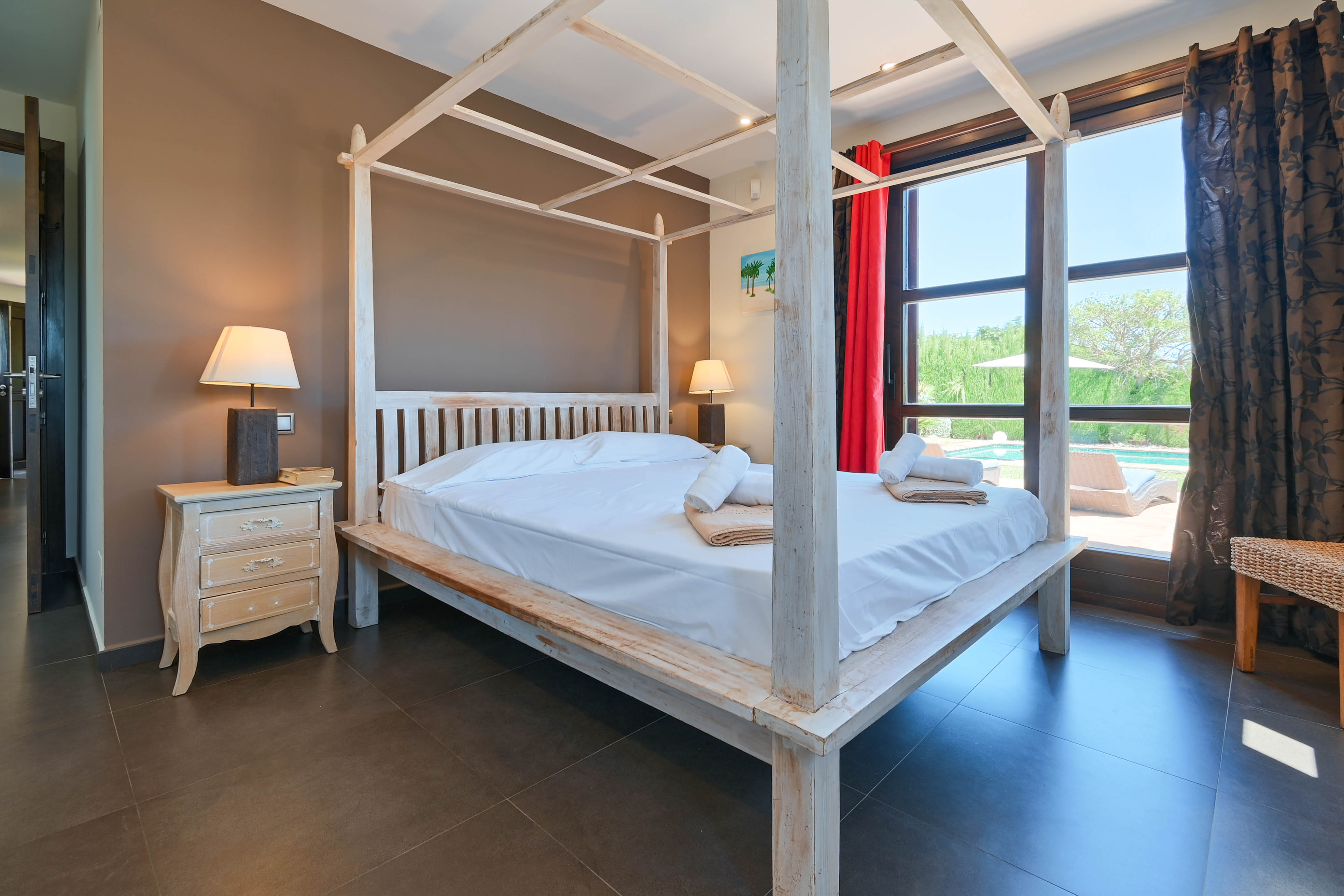 Chambre avec lit à baldaquin en bois blanc, table de chevet patinée et vue sur la piscine.