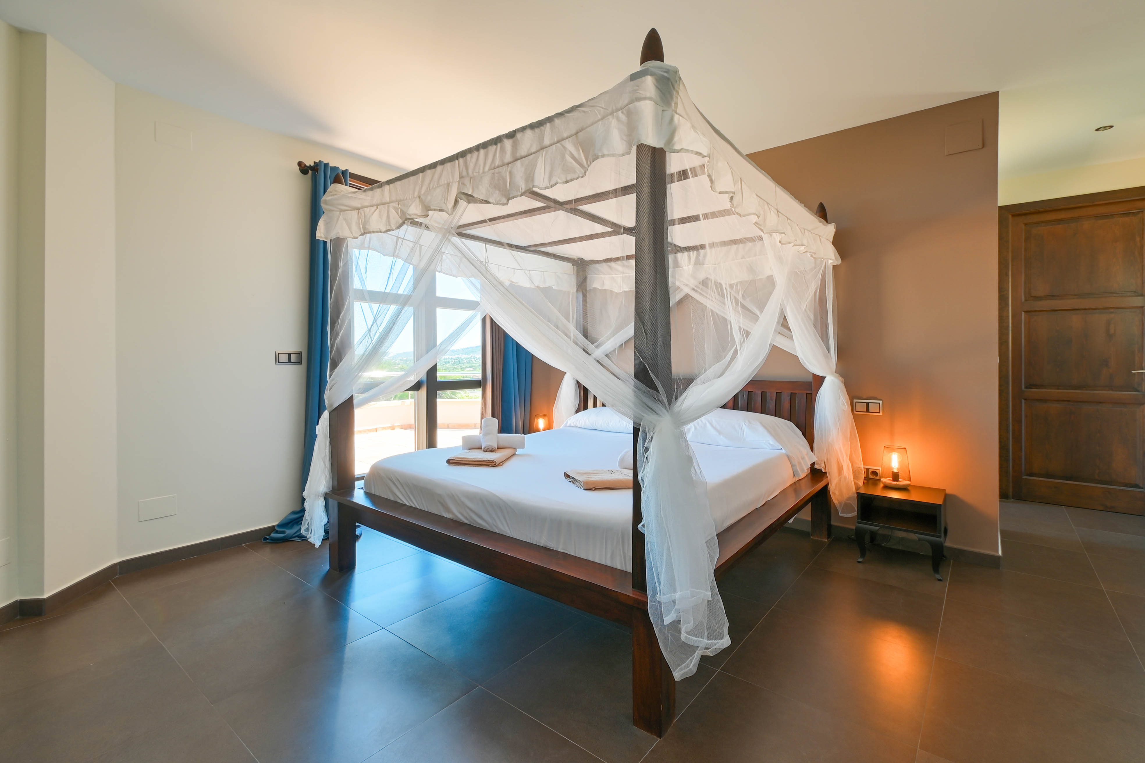 Chambre avec lit à baldaquin en bois foncé et rideaux blancs, sol carrelé marron.