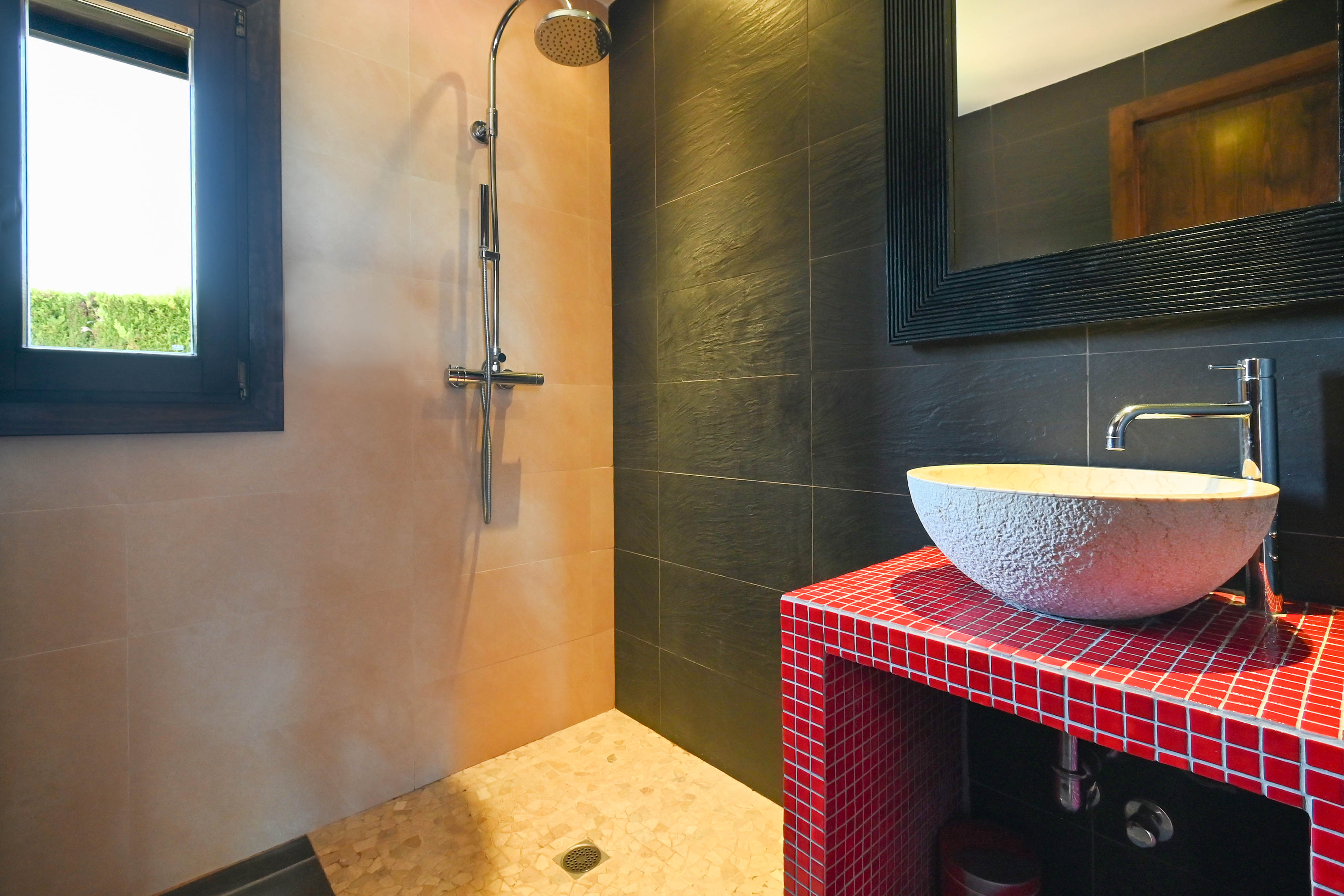 Détail de salle de bain avec lavabo rustique sur comptoir en mosaïque rouge et douche avec carrelage couleur sable.