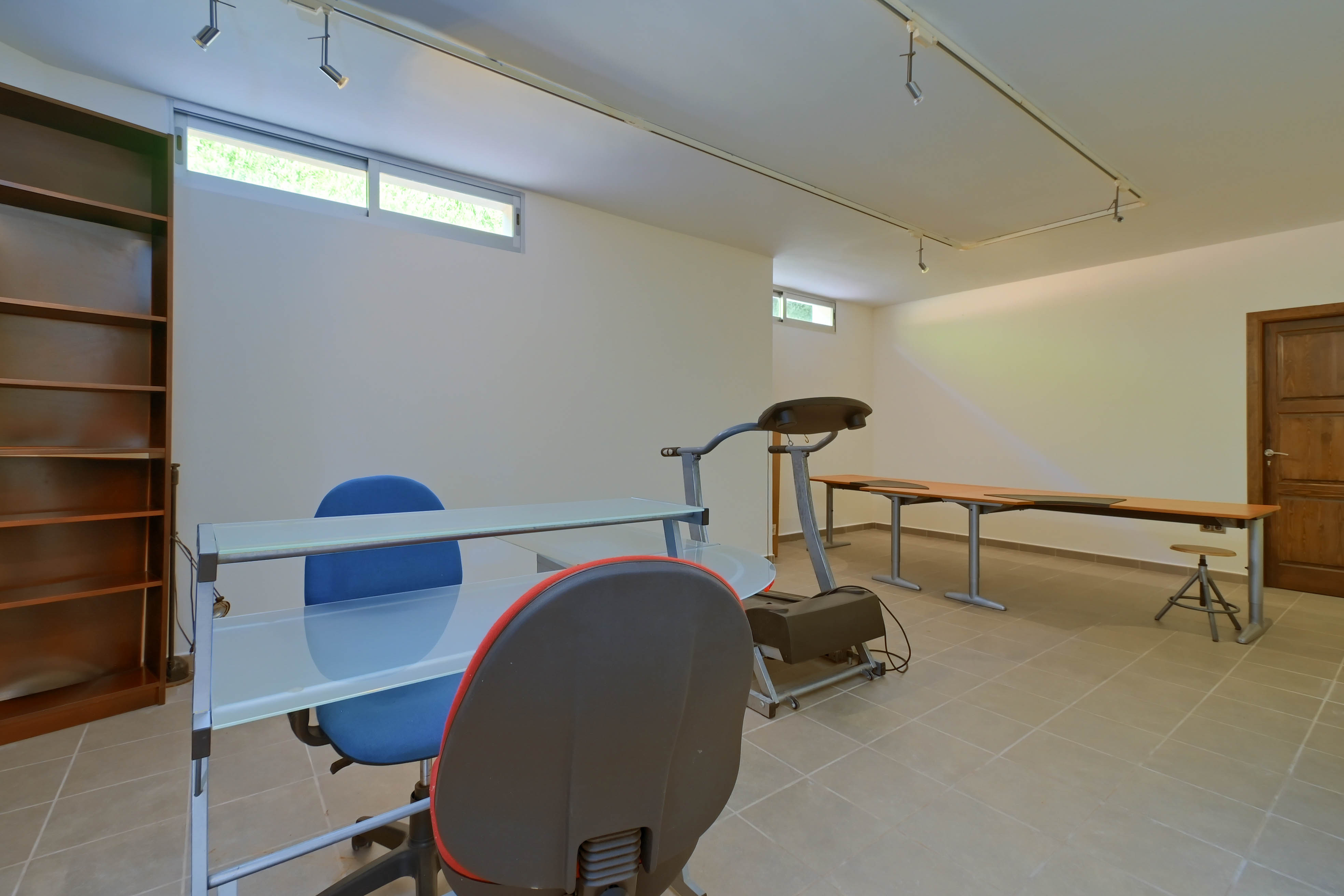 Espace fonctionnel avec étagères, bureau en verre et tapis de course, sur carrelage neutre.