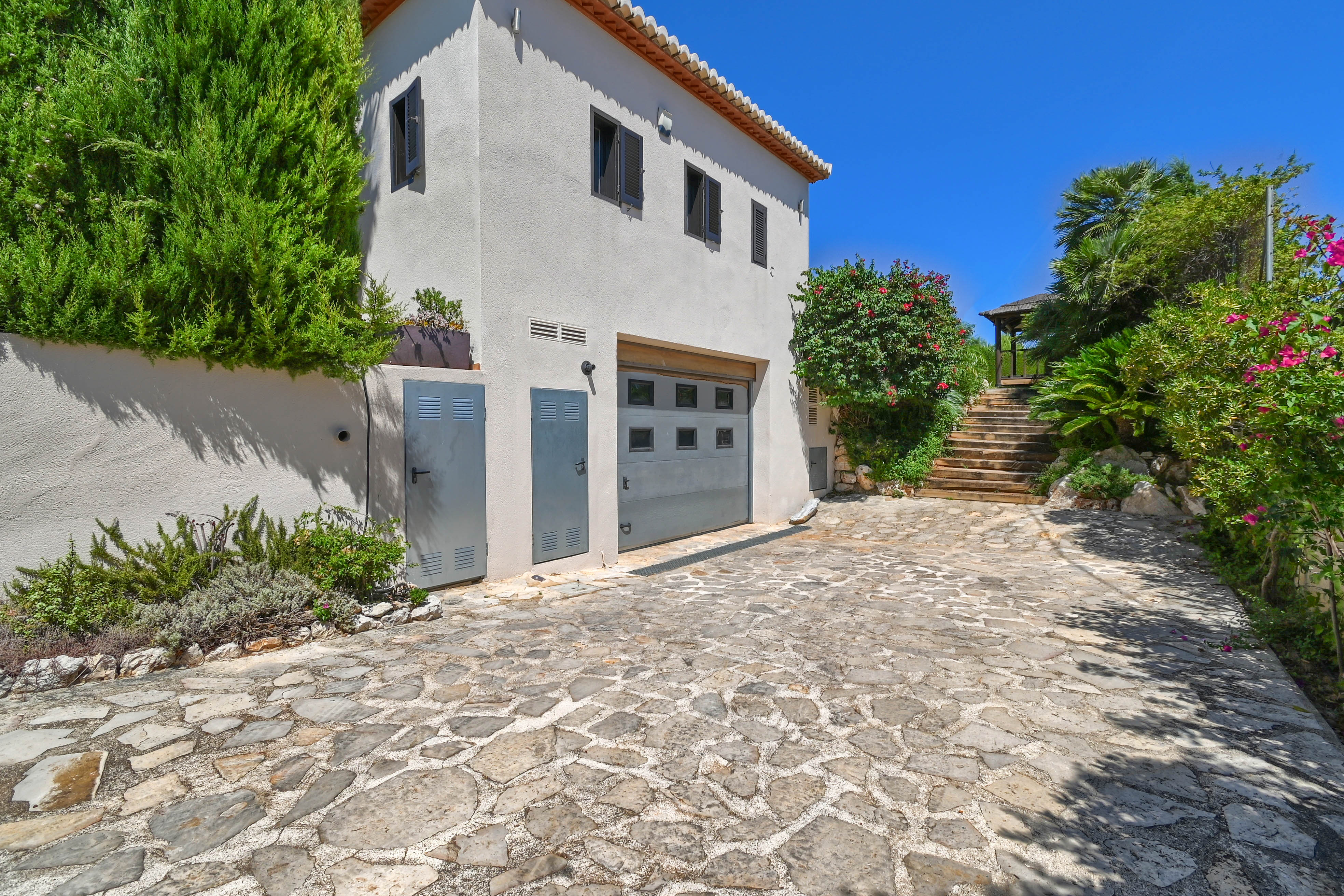 Extérieur de la villa avec allée en pierre, verdure et garage gris.